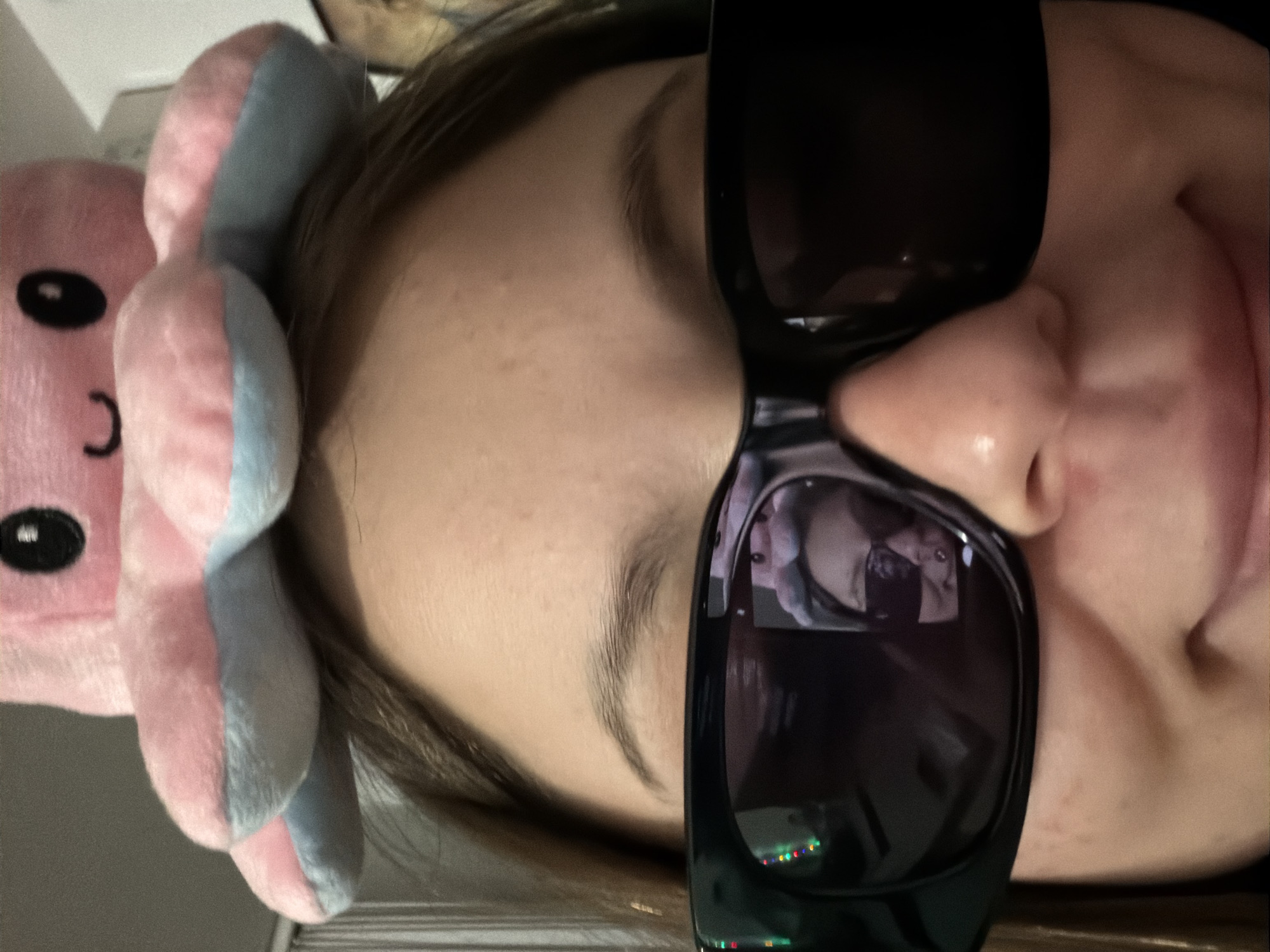 A person with brown hair is wearing black sunglasses and a pink and blue octopus headband. The person's face is turned to the side. The reflection in the sunglasses shows a room with a person and colorful lights. The location is not determinable from the image.
