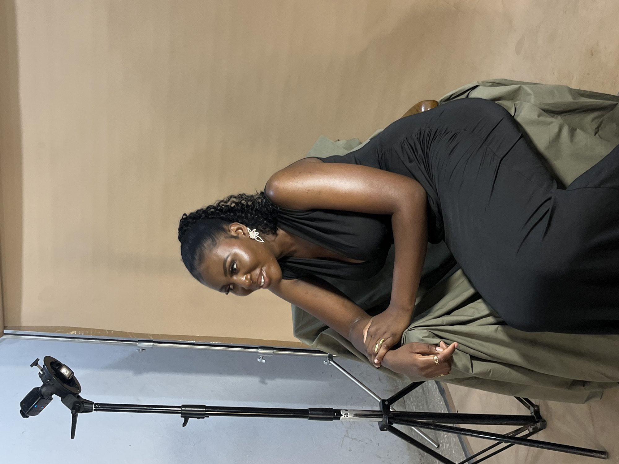 A woman in a black dress is posed on a chair, her body angled towards the camera. Her hair is pulled back, and she is wearing large, sparkly earrings and rings on her fingers. The background is a plain, light brown color, and to the left of the frame, a lighting stand with a flash is visible. The woman appears to be smiling and looking down, possibly at something just out of frame. The scene suggests a studio setting for a photoshoot. The lighting is soft, and there are no strong shadows. The overall mood is relaxed and confident. The location is specified as Gura Top, Nigeria. The time of day and weather are not discernible from the image. No text is visible in the image.