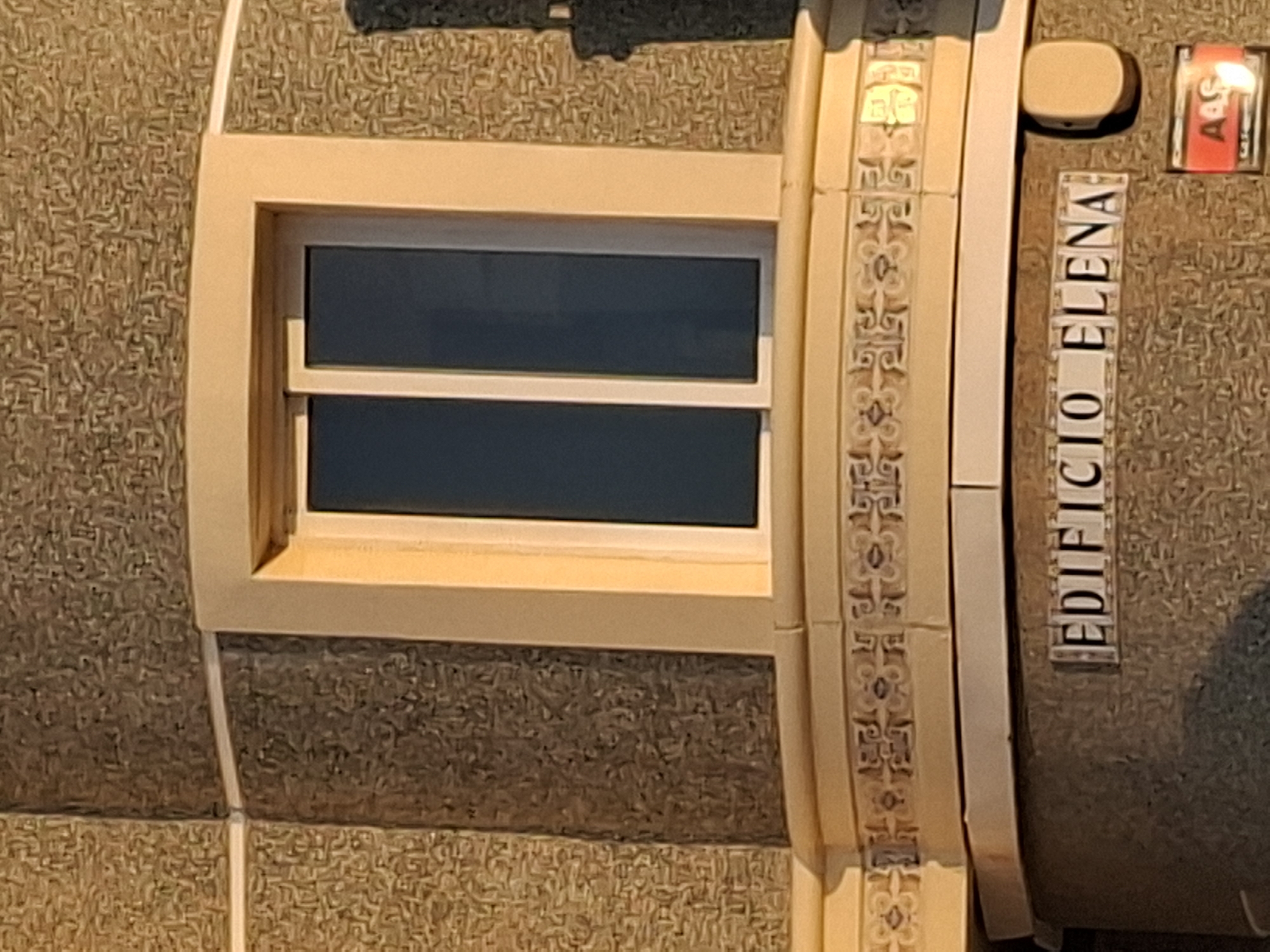 The image shows a close-up of the exterior of a building with a textured facade. A large window with white frames and dark glass is visible, recessed into the wall. Below the window, there is a decorative band of tiles with a repeating pattern. At the bottom of the image, a plaque reads "EDIFICIO ELENA" in white lettering on a dark background, indicating the name of the building. To the right of the plaque, a small red sign with white lettering appears to be an alarm or security system indicator. The lighting suggests it is either early morning or late afternoon, casting shadows on the building. This building is located in Torrevieja, Spain.