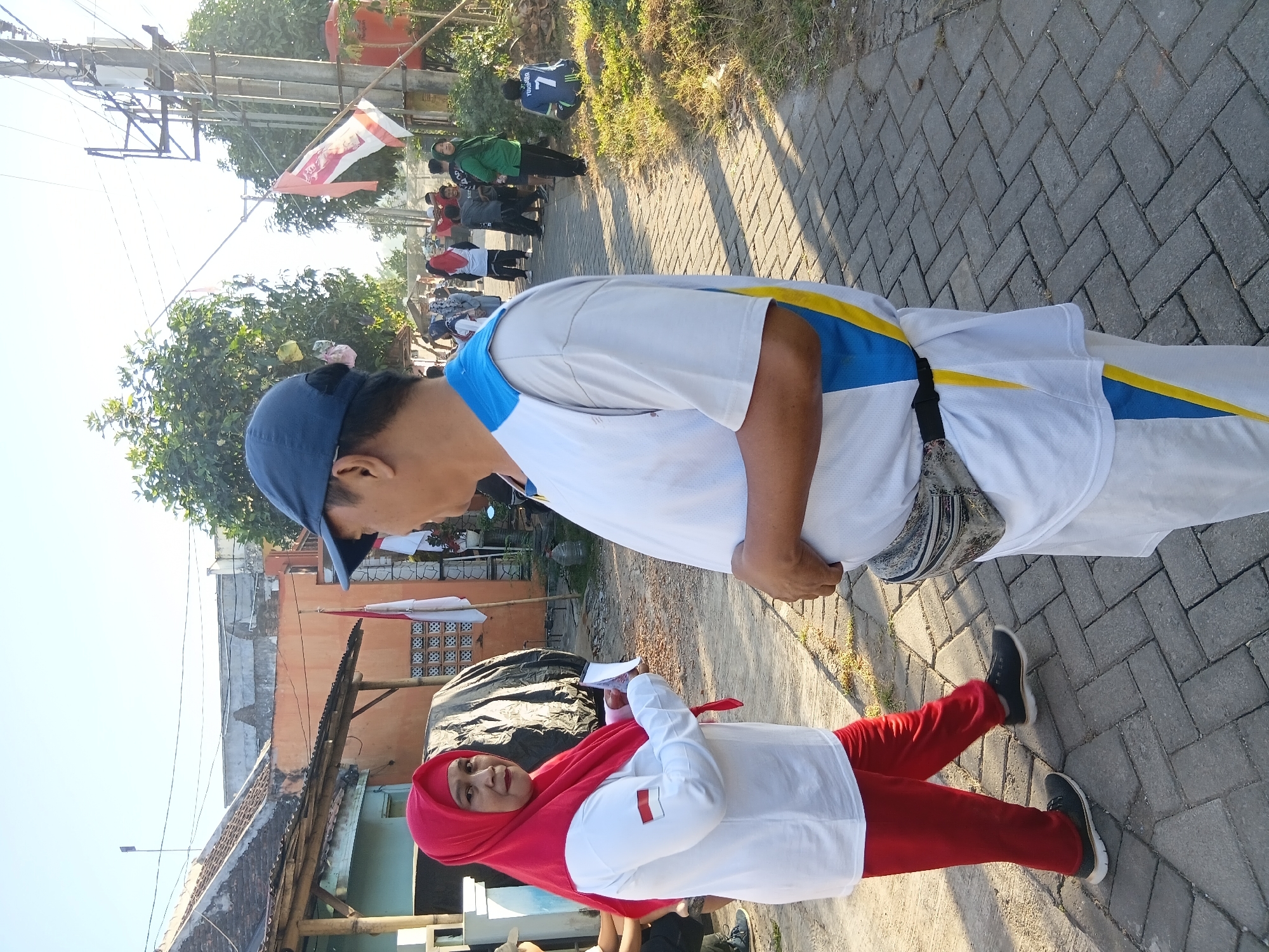 In Tanggul Wetan, Indonesia, a woman wearing a red hijab and a white long-sleeved shirt with a red and white flag emblem on the left sleeve, and red pants, stands on a brick-paved street, holding papers. She is looking at a man in a blue baseball cap and a white shirt with blue and yellow accents, who is facing her. The man has a fanny pack around his waist. In the background, other people are walking and gathered, with Indonesian flags visible on poles. The scene appears to be an outdoor event or gathering in a residential area, with houses and trees lining the street. The lighting suggests it is daytime, possibly morning or late afternoon.