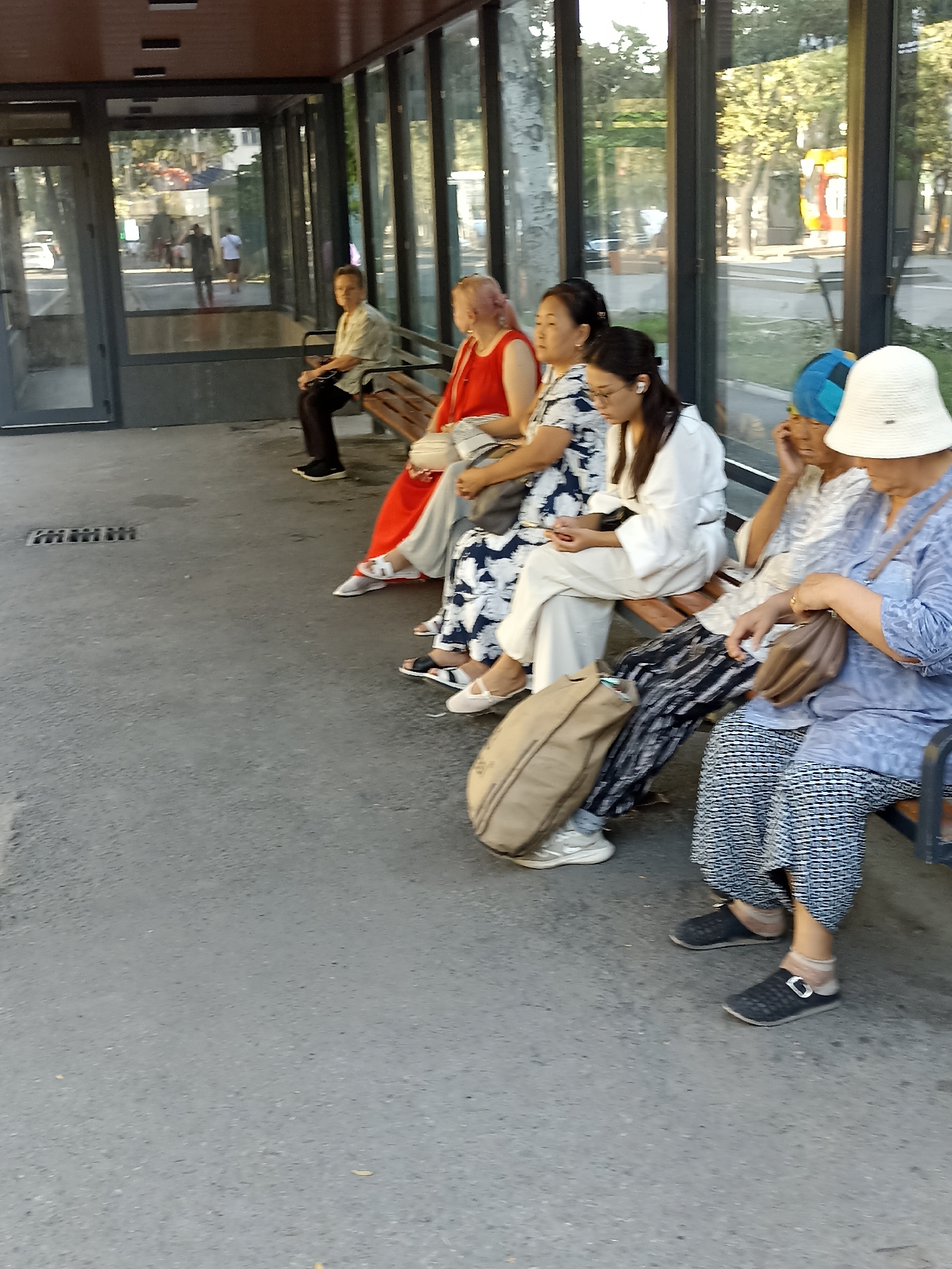 The image depicts a group of people sitting on a bench in a sheltered outdoor waiting area, likely a bus stop or a similar public transportation hub. The setting appears to be urban, with a paved ground and a glass-enclosed structure behind the bench. Trees and buildings are visible in the background through the glass, suggesting a city environment. The people are diverse in age and gender, all dressed in casual attire. Some are looking at their phones, while others are engaged in conversation or simply waiting. Bags and personal belongings are placed near the individuals, indicating they are either traveling or spending time outdoors. The overall atmosphere is relaxed and ordinary, capturing a moment of everyday life in a public space. Without specific landmarks or signs, the exact location in Bishkek City, Kyrgyzstan, cannot be definitively identified.