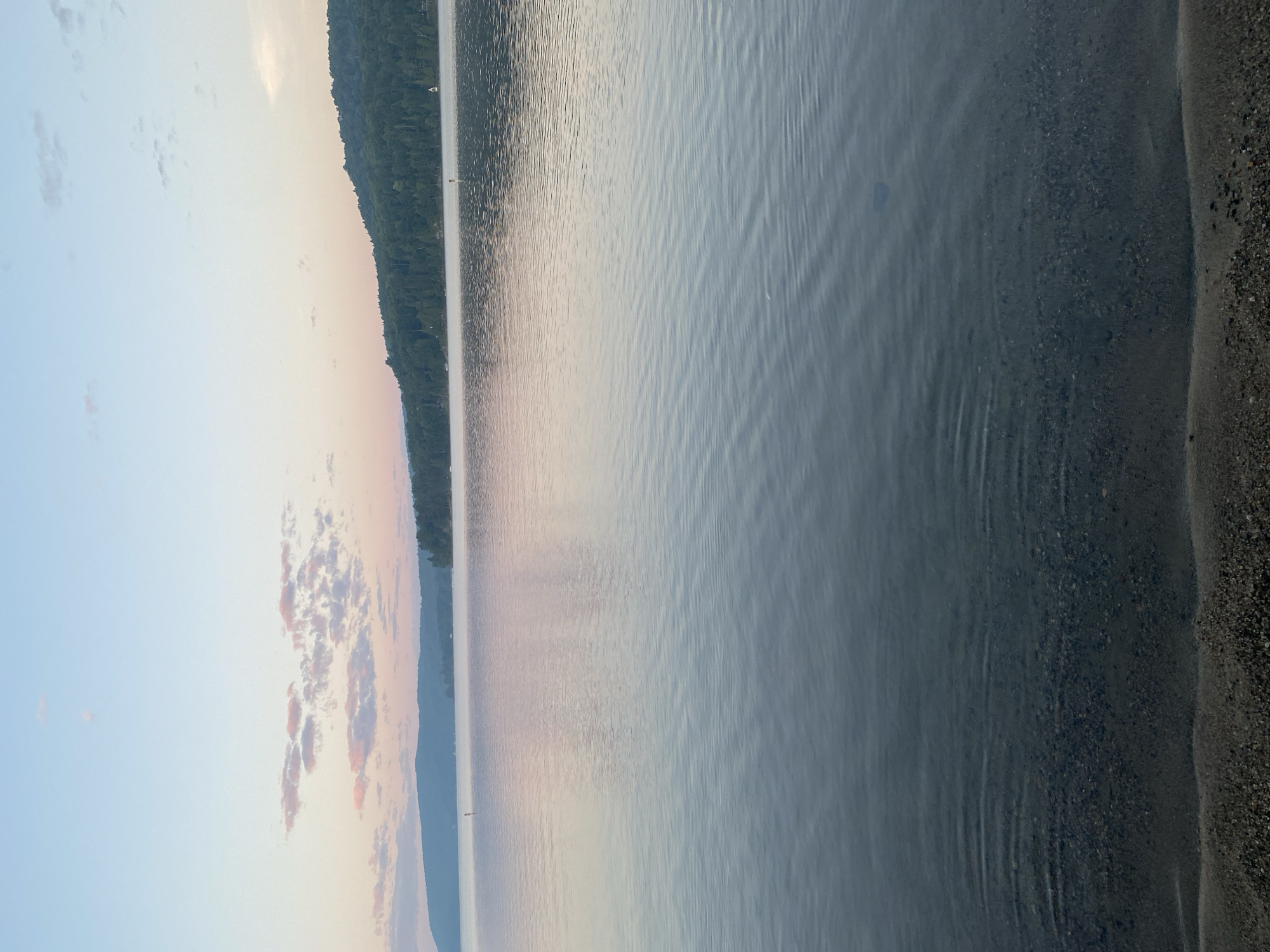 The image depicts a serene lake scene at sunrise or sunset. The sky is a soft gradient of blue and pink, with scattered wispy clouds. A forested hill forms the horizon line across the lake. The water is calm with gentle ripples, reflecting the colors of the sky. In the foreground, the edge of a pebble beach is visible. The scene appears to be located on the shores of Lac Simon in Duhamel, Quebec, Canada, judging by the characteristic landscape and the likely geographical context of the prompt.