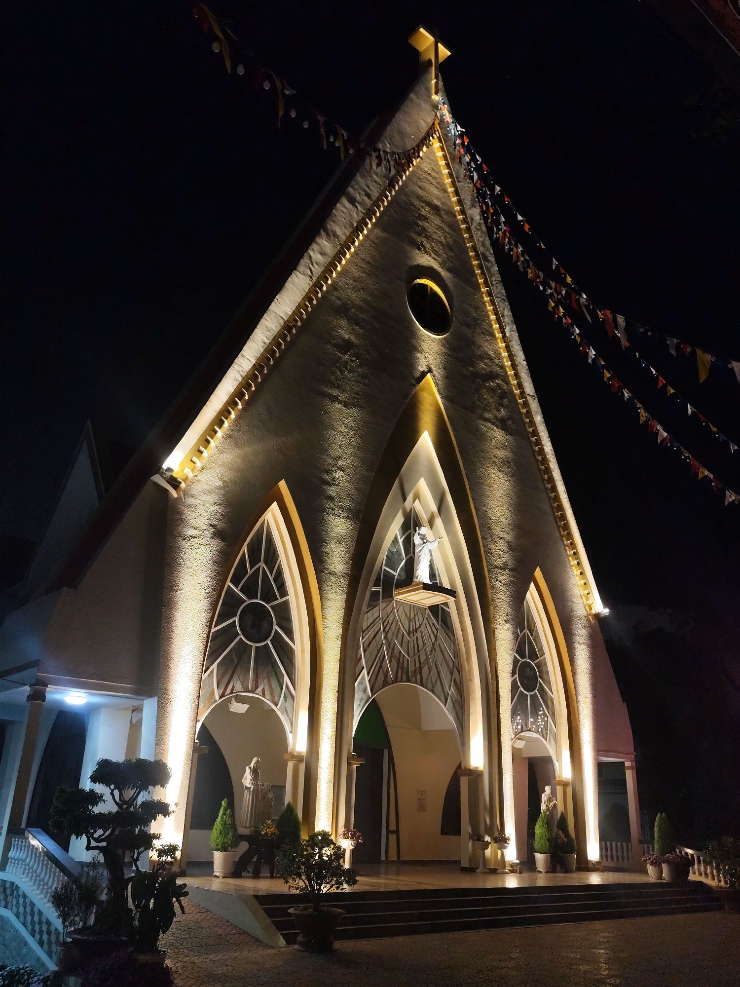The image depicts a church at night in Da Lat, Vietnam. The church's facade is brightly lit with golden lights accentuating its architectural features. The structure has a triangular roof topped with a cross, decorative flags hanging from the roofline, and three arched windows above the entrance. Statues are visible in the center window and flanking entrances. Potted plants and trees decorate the entrance area. The foreground shows a paved area leading to the church steps.