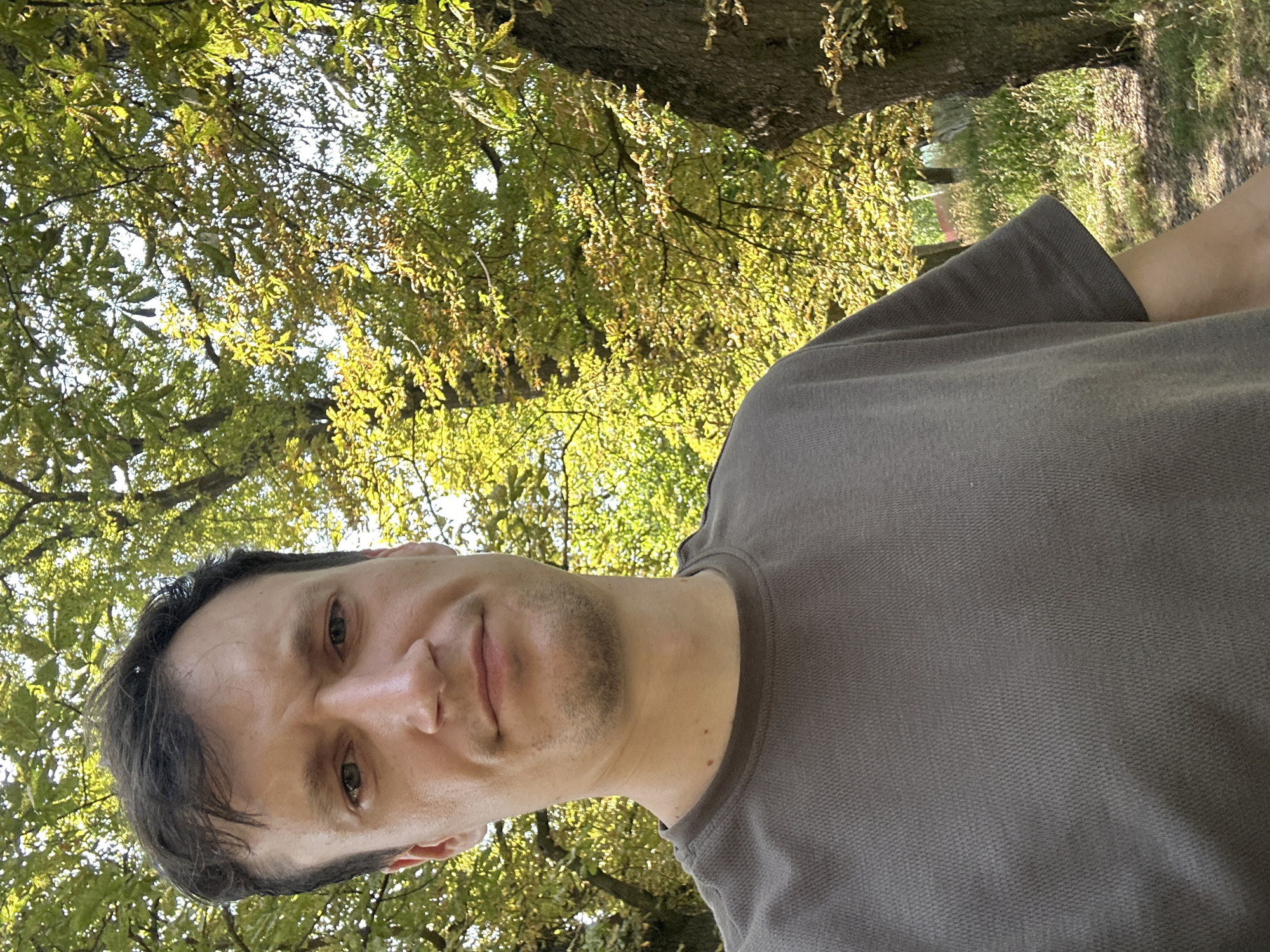 In Makiv, Ukraine, a fair-skinned male with short brown hair is captured in a selfie taken outdoors. He is wearing a dark grey t-shirt and is positioned in the foreground, with a backdrop of lush green trees and dappled sunlight. The trees, some of which appear to be chestnut, exhibit vibrant green foliage, suggesting a warm season. A large tree trunk is visible to the right of the man, and a dirt path winds through the grassy area behind him. The overall impression is that of a casual outdoor scene, possibly a park or wooded area.