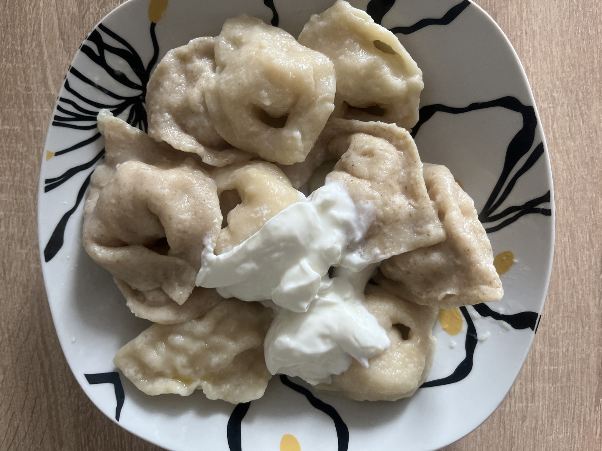 A close-up, overhead shot shows a white plate with a black and yellow abstract pattern. The plate is filled with dumplings, which appear to be a pale brown color. A dollop of white sour cream is placed on top of the dumplings. The plate is situated on a light-colored wooden surface. The image is a food photograph taken in a domestic setting, likely a kitchen or dining area, in the city of Rybnitsa, Moldova.