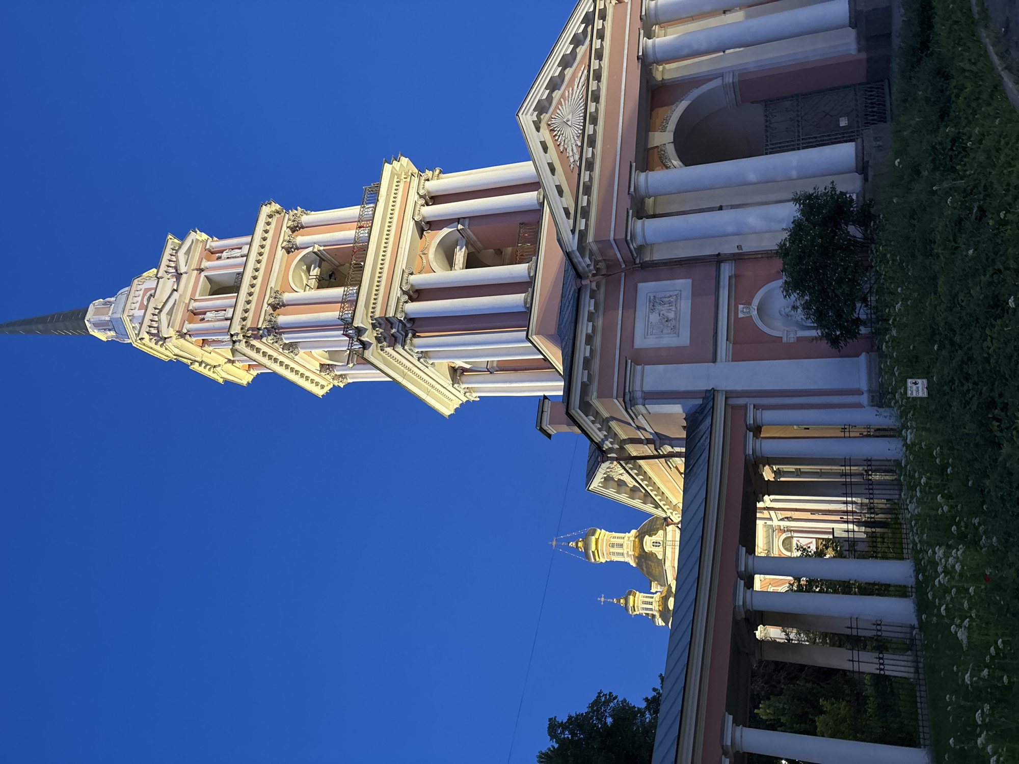 The image depicts the Trinity Cathedral, also known as the Izmaylovsky Cathedral, located in Saint Petersburg, Russia. The photograph, taken at dusk or night, showcases the cathedral's prominent pink and white belfry, illuminated by artificial lights. The belfry features multiple tiers with classical columns, arched openings for bells, and a tall, slender spire. To the left of the belfry, the golden domes of the cathedral's church are visible against the deep blue sky. In the foreground, a colonnade with white columns creates a covered walkway, leading towards the main building. A low fence separates the pathway from a grassy area with flowering plants and bushes. The overall scene is serene, highlighting the architectural grandeur of the cathedral in the twilight.
