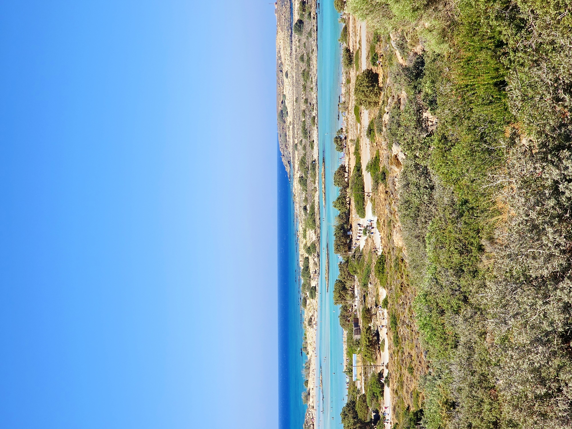 This is a vertical, eye-level, medium shot photograph of a scenic coastal landscape under a clear, bright blue sky. In the foreground, dense green shrubbery covers the lower portion of the image, gradually transitioning to sparser, dry, brush-like vegetation with scattered rocks and a few olive trees as it recedes towards the middle ground.
The middle ground features a shallow, turquoise lagoon with sandy shores and several small islands covered in sparse vegetation. Numerous people are visible, some wading in the shallow water, others walking along the shore or on pathways. A few small structures, including what appears to be a yellow building, are situated near the edge of the lagoon. A paved pathway or road runs through this area, with more people walking along it.
In the background, the lagoon opens up to the deeper blue sea, which stretches to the horizon. A rocky, arid peninsula with sparse vegetation extends into the sea on the right side of the background. The overall impression is a popular, sunny beach destination. The location is identified as Elafonisi Beach, Greece.