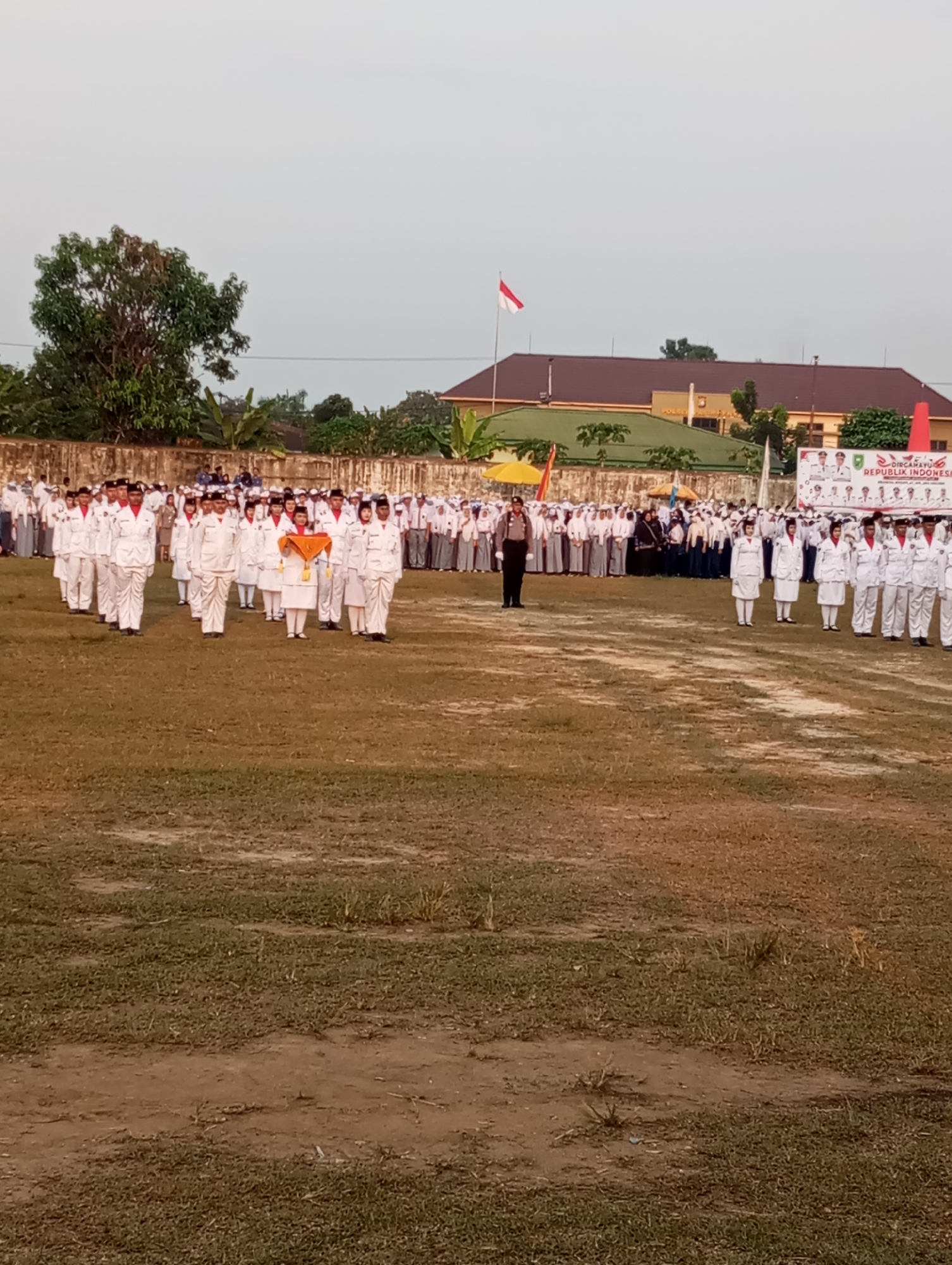 The image shows an outdoor event in Candirejo, Indonesia, likely a school or government ceremony. A large group of people, predominantly young students in white uniforms, are assembled on a grassy field. Many are standing in neat rows, appearing to be part of a formal procession or parade. Some individuals in the front are holding flags or banners, including one prominent orange and yellow banner. A few adults in uniform, including a police officer, are also present and appear to be supervising or leading the event. In the background, there are buildings with a reddish-brown roof, a wall, and trees. The sky is overcast.