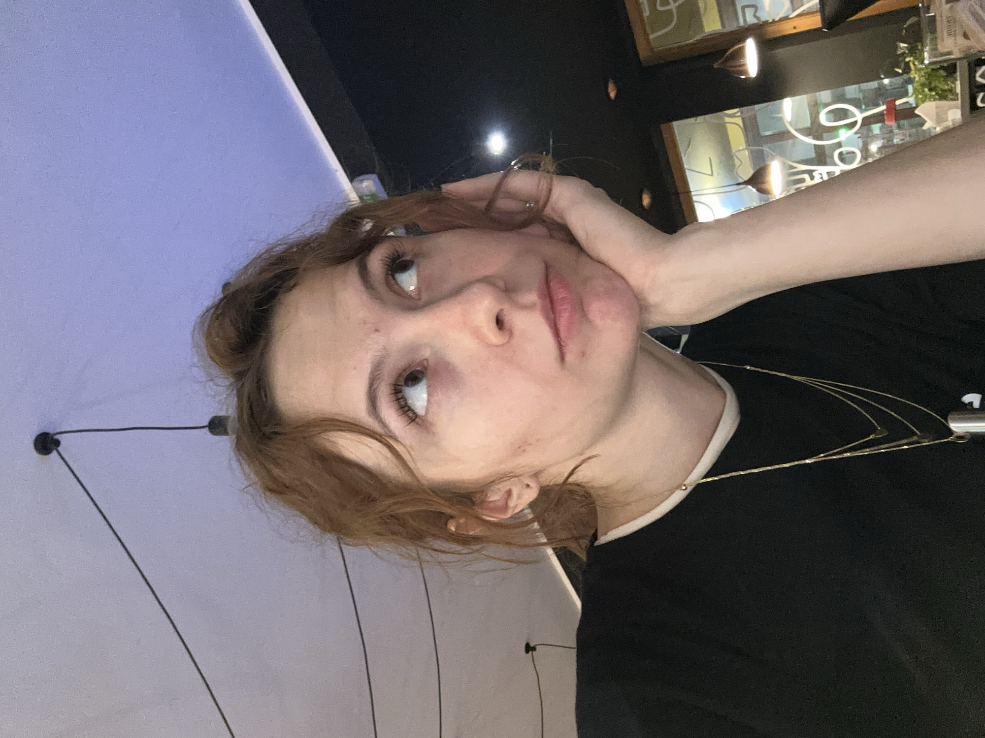 A woman is posing for a selfie. She has light skin, reddish-brown hair, and is wearing a black t-shirt and multiple gold necklaces. Her head is tilted, and her hand is resting on her cheek. The background consists of a mix of elements. To the left, there is a light purple wall with a light fixture attached to it. To the right, there is a darker area with what appears to be a window showing an illuminated sign or decoration, and hanging light fixtures. Based on the neon sign visible through the window, the location is Novi Sad, Serbia.