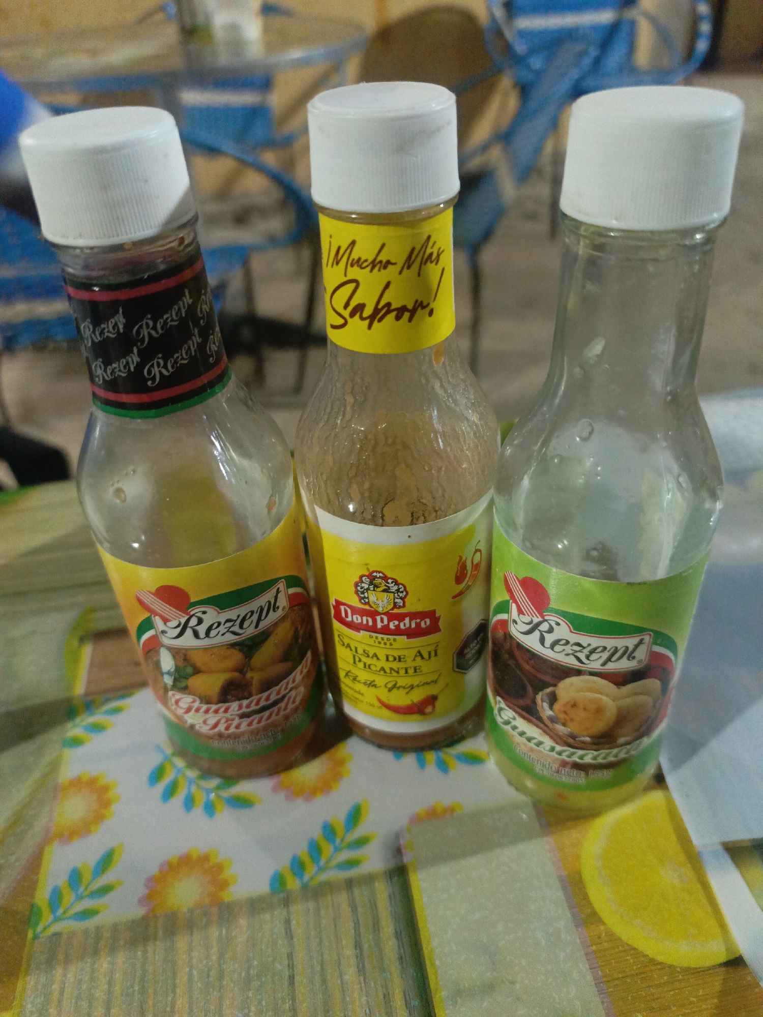 The image shows three bottles of hot sauce standing on a table with a patterned surface, likely a tablecloth. The bottles are all partially empty. From left to right, the first bottle has a black and red label with "Rezept" printed on it, over a yellow label with "Guasacaca" also printed on it. The middle bottle is yellow, with "Don Pedro" and "Mucho Más Sabor!" written on it, along with images of chili peppers. The third bottle has a green label with "Rezept" and "Guasacaca" on it. The bottles all have white caps. In the background are blue chairs. The location may be La Tagua, Venezuela, based on potential associations of the brands or styles of products depicted, but without further information, this cannot be confirmed.