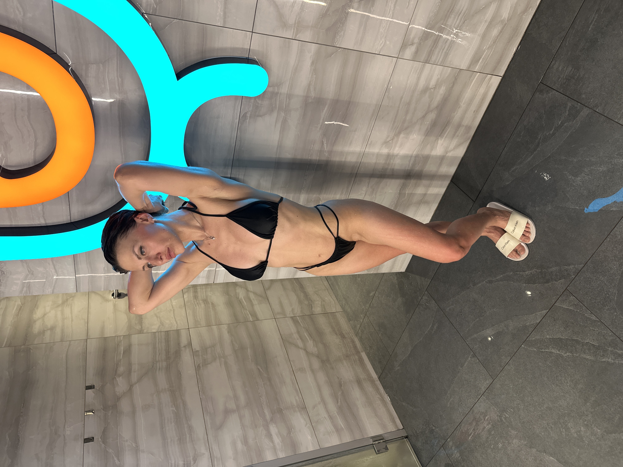A woman in a black bikini and white Calvin Klein branded sandals is lying on her side on the floor of a modern tiled room. The walls and floor have a gray tile pattern, with different shades creating contrasting sections. Above her is a bright orange and cyan sign, possibly an illuminated design element. The setting appears to be indoors, possibly in a sauna or spa area, judging by the tiles and shower fixture in the background. There is no explicit information in the photo to indicate the exact location in Saint Petersburg, Russia.