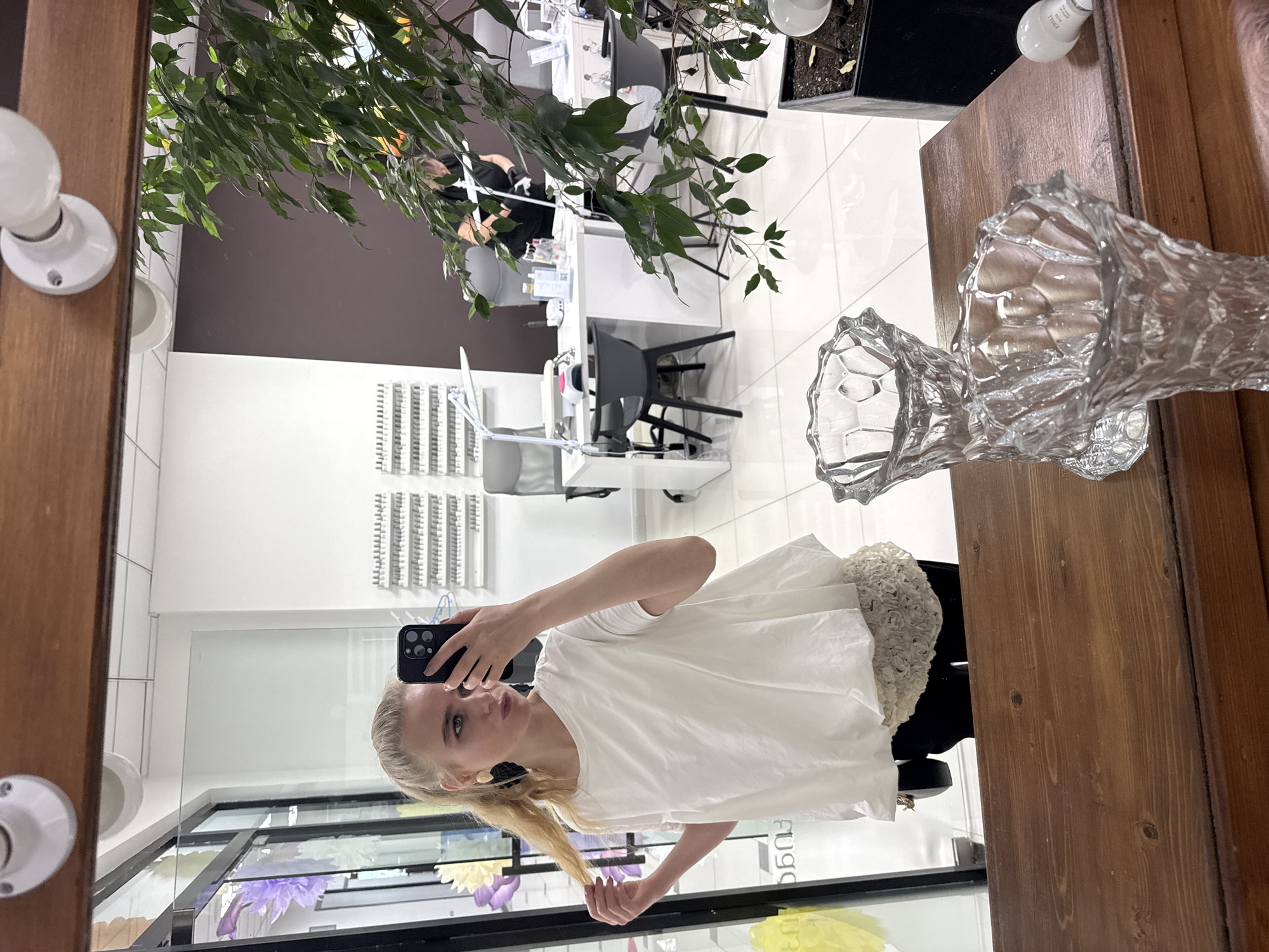 A blonde woman in a white top takes a selfie in a mirror. The reflection shows a salon interior with manicure stations, chairs, and nail polish displays. The city is Unknown, Unknown. A crystal vase and green plant are next to the mirror on a wooden surface.