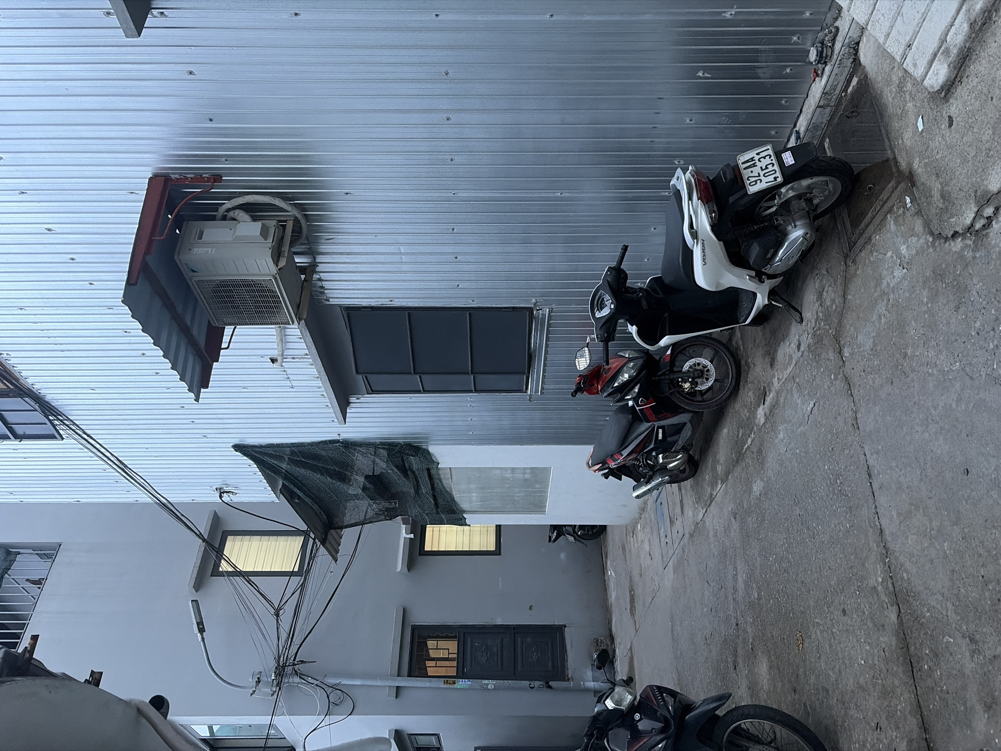 The image depicts a narrow alleyway in Da Nang, Vietnam, flanked by two buildings. The building on the right is constructed with corrugated metal siding and features a mounted air conditioning unit under a red and silver awning. A dark-paned window is visible on this building. The building on the left is made of concrete, painted a light gray, and has at least two windows with blinds. A street lamp is attached to this building, with a dense network of electrical wires extending from it. There are several parked motorcycles in the alley: a black motorcycle on the left, a red and black motorcycle in the middle, and a white and black motorcycle on the right. The ground is paved with concrete, showing some cracks and puddles. One of the motorcycles has a license plate that reads "92-AA 405.31". A dark mesh tarp is draped over a section of the left building.