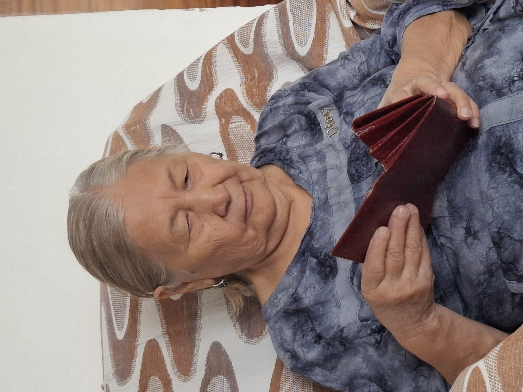 An elderly woman with grey hair and wrinkles on her face is sitting in a chair and looking at a red wallet. She is wearing a blue, patterned shirt. The chair has a brown and white patterned fabric. The background is a plain white wall. The image is a close-up shot, focusing on the woman and the wallet. The scene appears to be indoors, possibly in a home. There is no indication of the specific location within the city of Almaty, Kazakhstan.