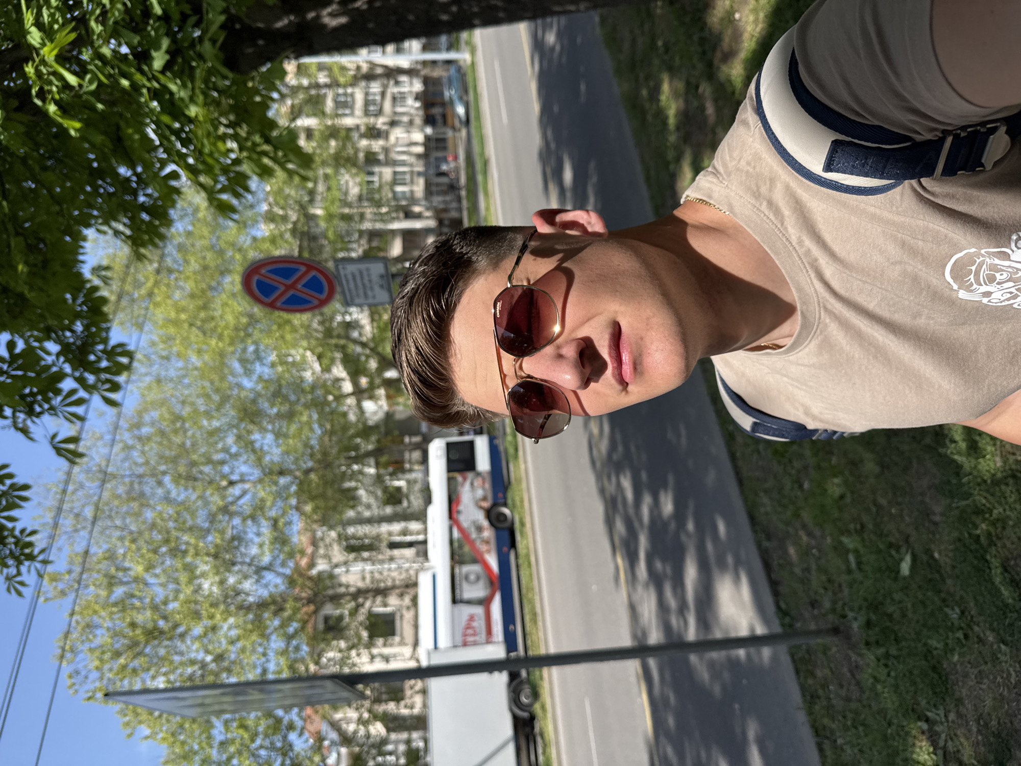 The image shows a man wearing sunglasses and a light brown t-shirt with a white design on it. He also wears a satchel with a white and blue strap over his shoulder. The photo appears to be taken at street level, near a sidewalk and a road. In the background, there is a street with buildings and a bus, a "No Stopping" sign is visible. Based on these details, it's likely that the photo was taken near a street with public transportation in a populated area. Without further information, determining the exact location in Burgas is not possible.