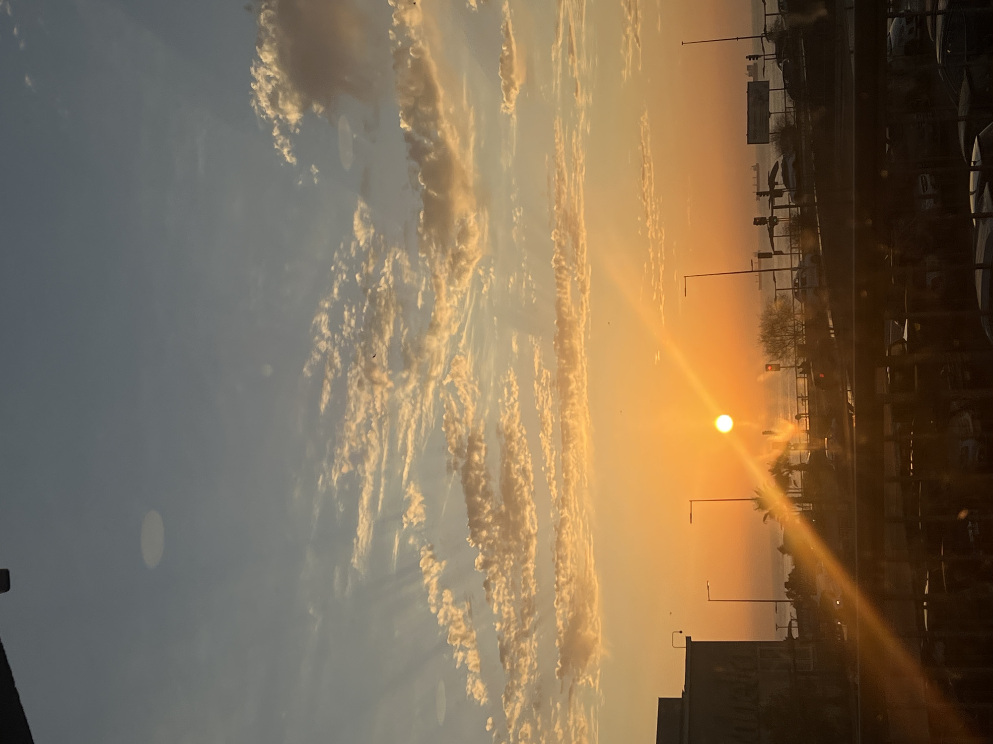 The image shows a sunset in Arica, Chile, with a view overlooking the Pacific Ocean. The sky is filled with clouds illuminated by the setting sun, casting a warm orange and yellow glow across the scene. A line of buildings and streetlights is visible along the coastline. The image appears to be taken from an elevated position, possibly from inside a building or vehicle. The lighting conditions suggest the time of day is either dawn or dusk.