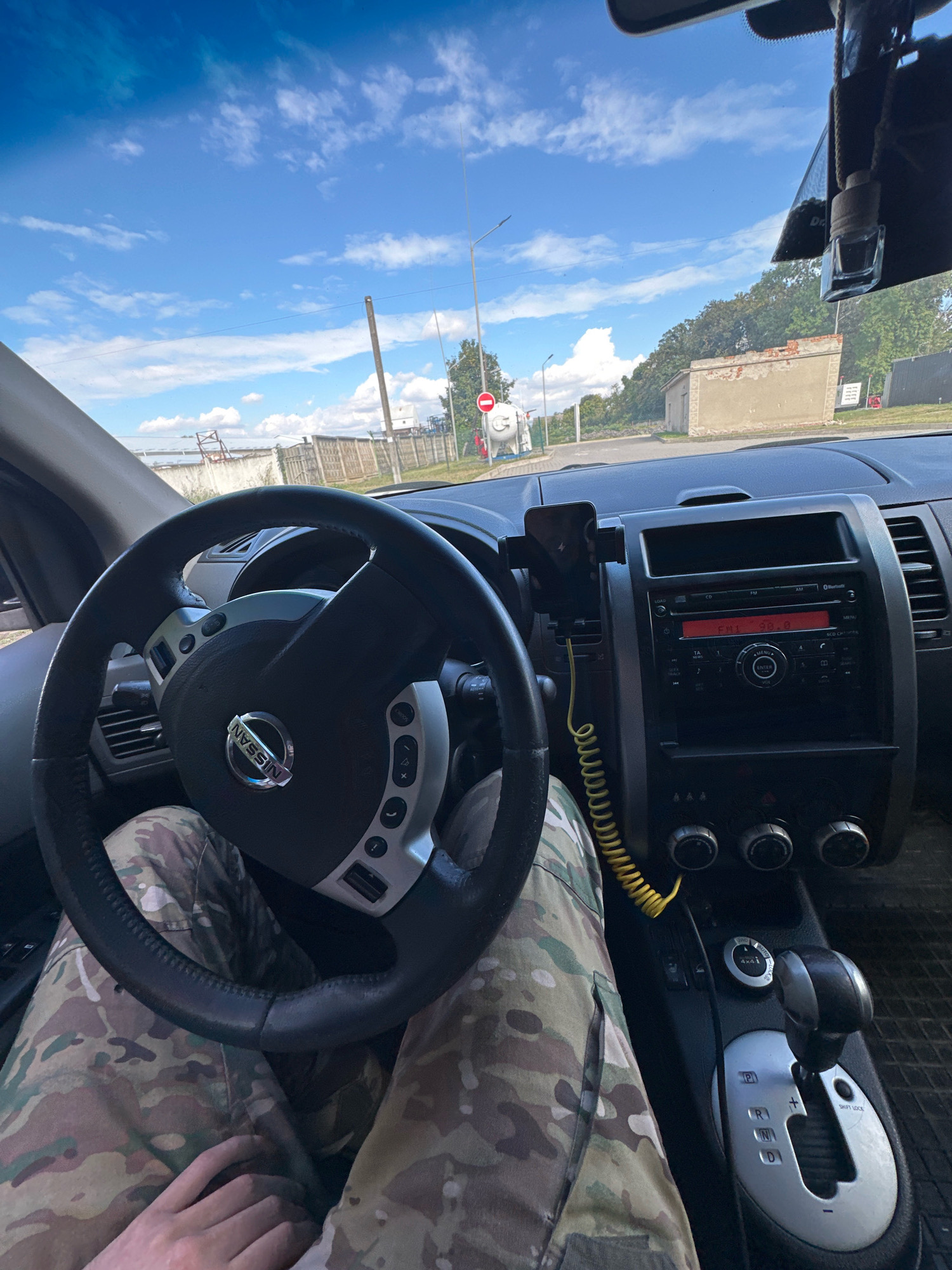 The image shows the interior of a car, likely a Nissan, parked outdoors. The viewpoint is from the driver's seat, looking forward. The steering wheel is visible on the left, with the Nissan logo in the center. The dashboard features a car stereo with a red screen displaying "FM1 89.0", climate control knobs, and a gear shift lever. A mobile phone is mounted on a black holder attached to the dashboard. The driver's legs, clad in camouflage pants, are visible in the foreground. Outside the car, a blue sky with scattered clouds is seen. There are trees and buildings in the background, including a brick structure and a large white industrial tank. A traffic sign with a red circle and a horizontal bar is also visible. The context of Shatava, Ukraine, is not directly evident from the visual information in the image itself.