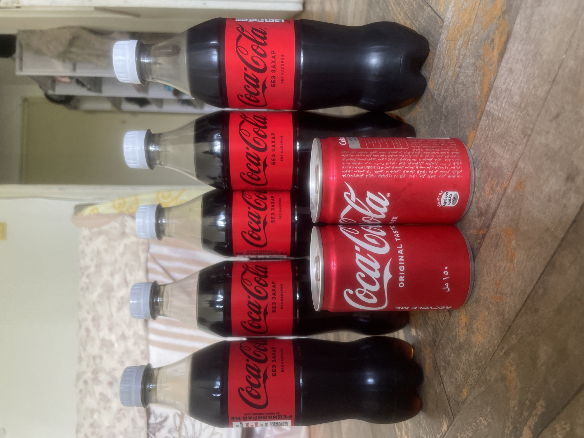 The image shows a collection of Coca-Cola products: five bottles labeled "Coca-Cola БЕЗ ЗАХАР БЕЗ КАЛОРИИ" (Coca-Cola Zero Sugar Zero Calories) and two Coca-Cola cans. The cans also have Arabic writing on them, with the text "ORIGINAL TASTE" and "RECYCLE ME". The products appear to be stacked on a wooden or wood-patterned surface. The bottles have white caps.
There is no indication of this image being in Plovdiv, Bulgaria, nor any information about people or interactions.