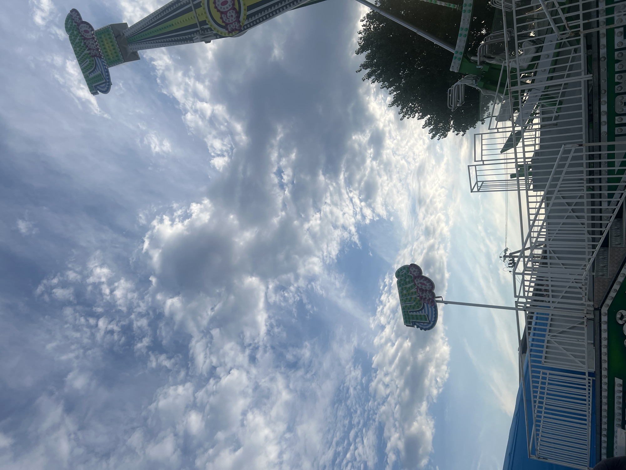 The image depicts a portion of a carnival ride, specifically the "Turbo" ride, under a cloudy sky in Peresyp, Russia. The ride is predominantly green and white and appears to be inactive. A tall, vertical structure of the ride, with a "Turbo" sign at the top, extends upwards. In the foreground, a white railing and stairs lead to the ride's platform, which is decorated with green and white lights. The sky is filled with scattered clouds, some of which are dark and heavy, while others are illuminated by sunlight, suggesting a dynamic weather pattern. A few trees are visible to the right of the ride. The overall scene is indicative of a fairground or amusement park. There are no people clearly visible in the frame.