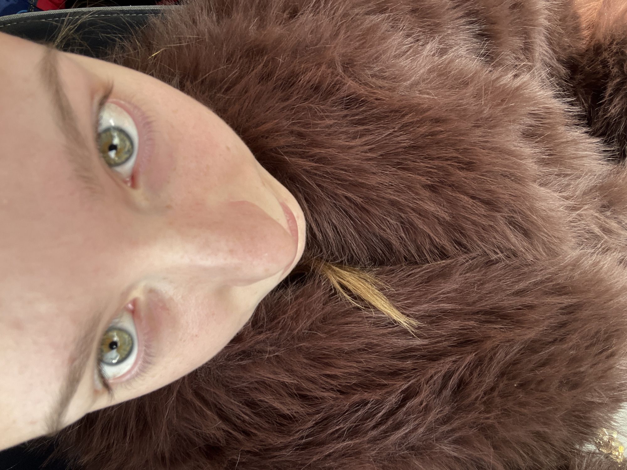 A close-up, angled portrait features a person with fair skin, freckles, and green eyes looking upward. The person is surrounded by a soft, brown, fur-like material. A few strands of light blonde hair are visible among the brown material. The location is not identifiable from this image, and there is no information to suggest it is in Varna, Bulgaria.