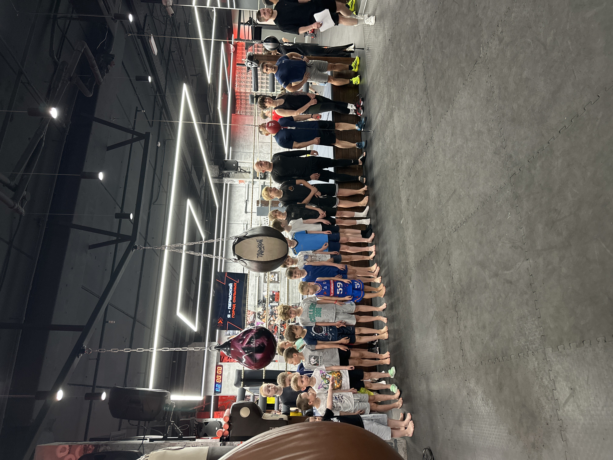 A slightly overexposed photograph shows a group of people lined up in a gymnasium. The floor is covered in black, interlocking mats. Hanging from the ceiling are multiple punching bags, one of which is a vibrant maroon color. A digital timer displays "88:88:00" in red digits. Neon lights in a zigzag pattern illuminate the scene. A sign visible in the background is in Russian, identifying the location as Perm, Russia. The group consists of approximately 20 individuals, primarily children, standing shoulder to shoulder. Behind the children stand several adults. Some individuals are barefoot, while others wear sneakers. Clothing varies, with some wearing athletic gear and others in casual attire. A man at the end of the line holds a paper. The orientation of the photograph is rotated, with the subjects appearing vertical.