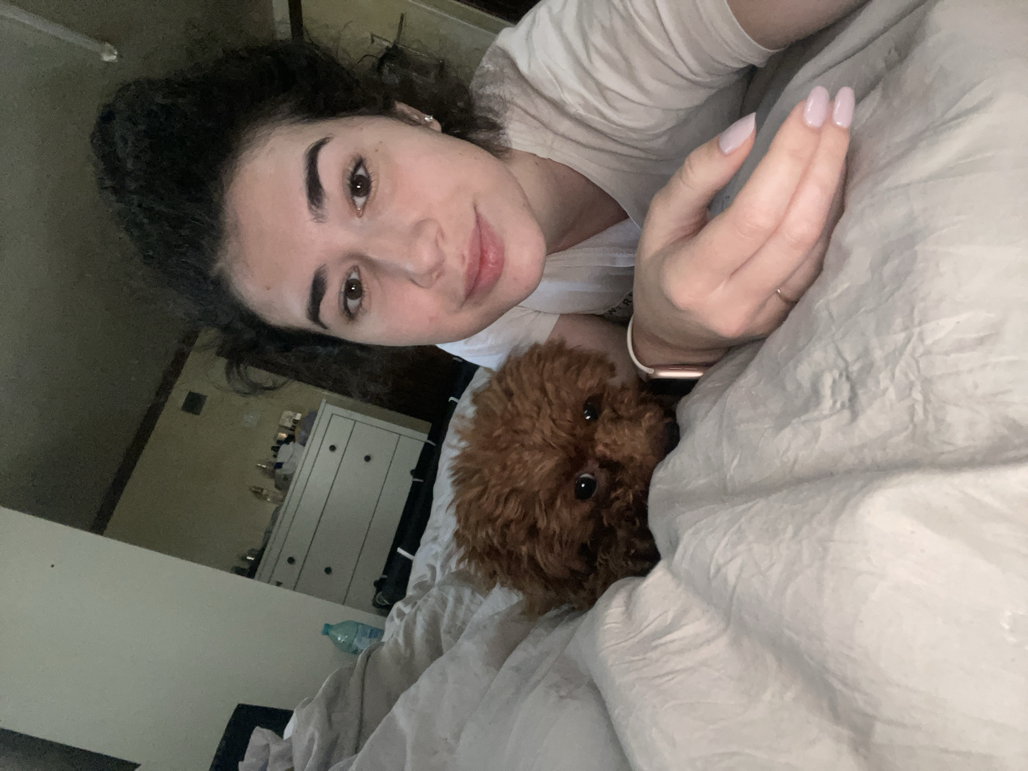 A woman is lying in a bed with light gray bedding, taking a selfie. She is wearing a white t-shirt and has dark curly hair and manicured nails. A small brown dog is partially visible, lying next to her in the bed. In the background, a white dresser is visible, with miscellaneous objects on top of it. A water bottle is on the floor near the dresser. The location cannot be determined to be Sofia, Bulgaria, based on the image.