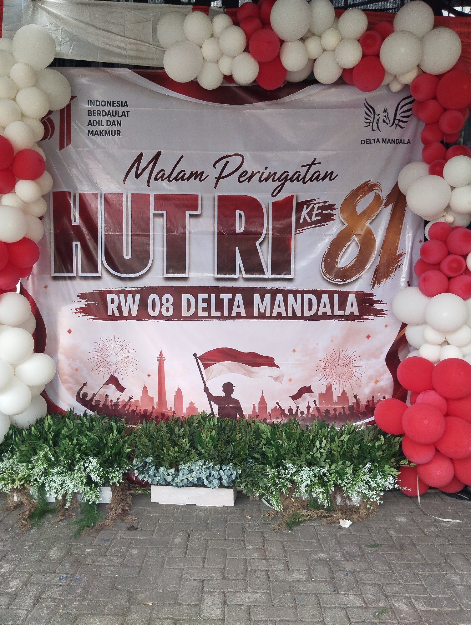 This is a photograph of a celebration banner in Semambung, Indonesia. The banner, decorated with red and white balloons, features the text "Malam Peringatan HUT RI KE 81" which translates to "81st Indonesian Independence Day Celebration Night." Below this, it reads "RW 08 DELTA MANDALA." The background of the banner depicts silhouettes of people celebrating with fireworks and the Monas tower, a landmark in Jakarta. In front of the banner, there are decorative plants and some scattered debris on a brick-paved ground.