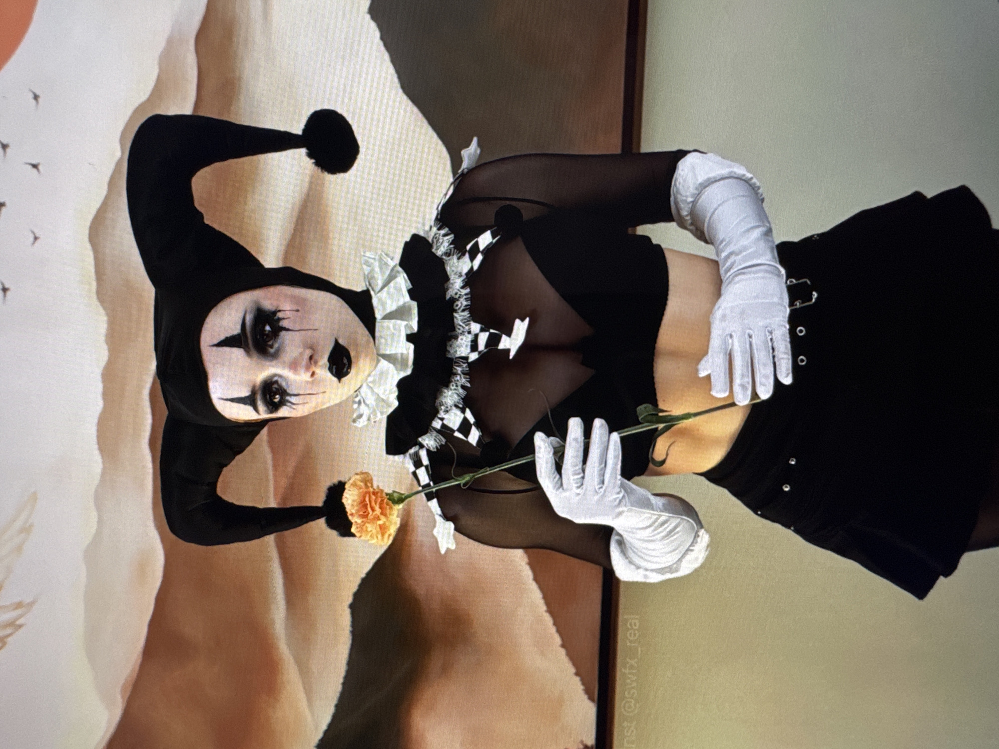 A person in a jester costume is shown from the chest up. The costume includes a black jester hat with two floppy points, a ruffled white and black checkered collar, and a black top with sheer sleeves. White opera-length gloves are worn on both hands. The person is holding a single orange carnation with their right hand. The makeup features dramatic dark eyeshadow and lipstick, with black teardrop-shaped markings under the eyes. The background is a blurred image of abstract mountain-like shapes in warm earth tones, with several small bird-like figures in the upper part of the frame. The lower left corner of the image contains a faint watermark that reads "inst @swfx_real". The lighting is soft and appears to be coming from the front, illuminating the subject and creating a shallow depth of field.