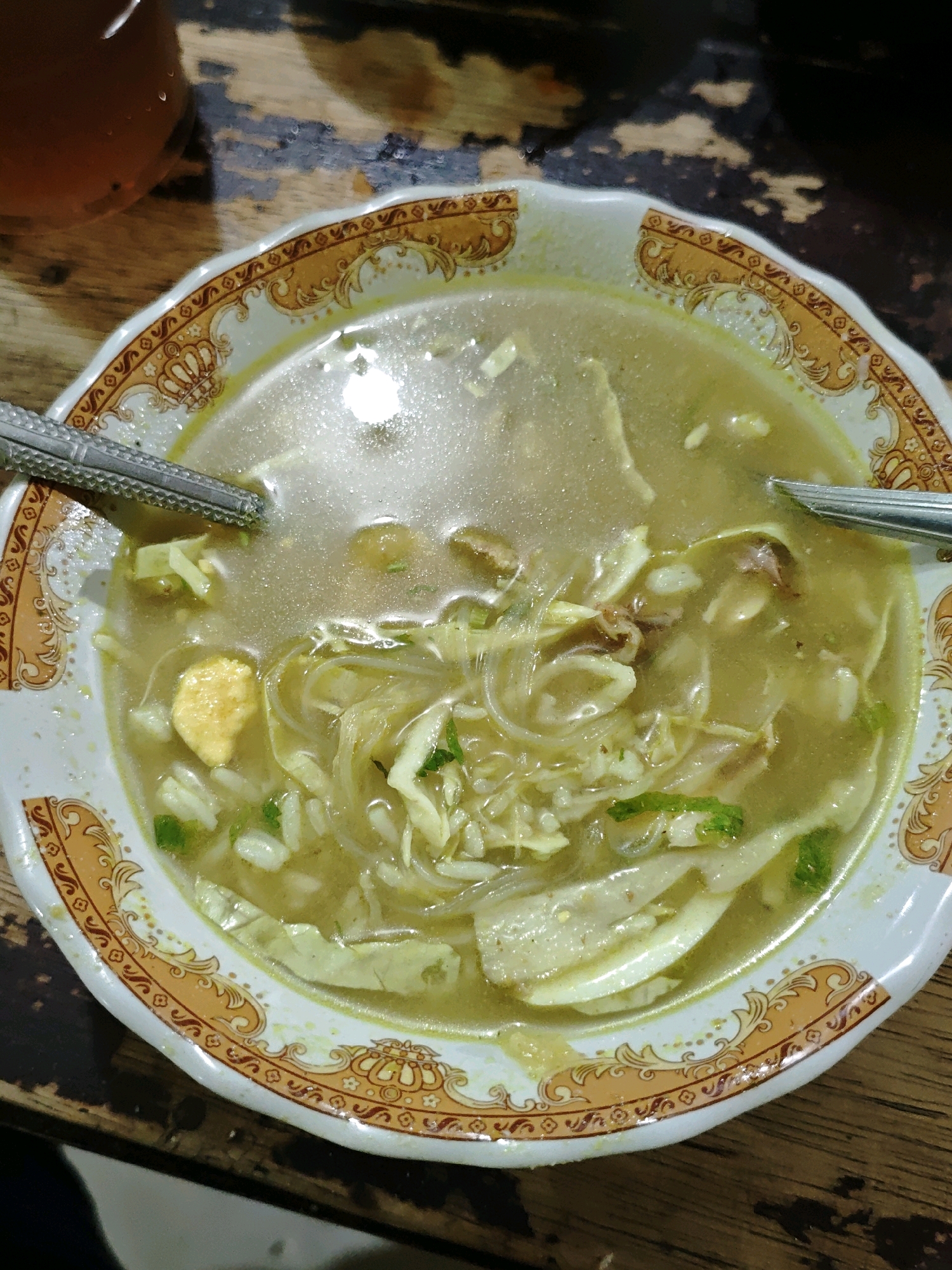 A close-up, overhead shot shows a bowl of soto ayam, a traditional Indonesian chicken soup, on a wooden table. The soup is served in a white ceramic bowl with an ornate orange and gold rim. Inside the bowl, thin strands of noodles, shredded chicken, and pieces of boiled egg are visible in a clear, golden broth. Sliced green onions are scattered throughout the soup, adding a touch of color. Two spoons, one on the left and one on the right, are placed in the bowl, suggesting the soup is ready to be eaten. The wooden table has a textured surface with visible grain and a dark, somewhat worn appearance. The overall lighting is dim, casting a soft glow on the food. The scene is set in a casual dining environment, likely a restaurant or home kitchen in Ngasem, Indonesia.