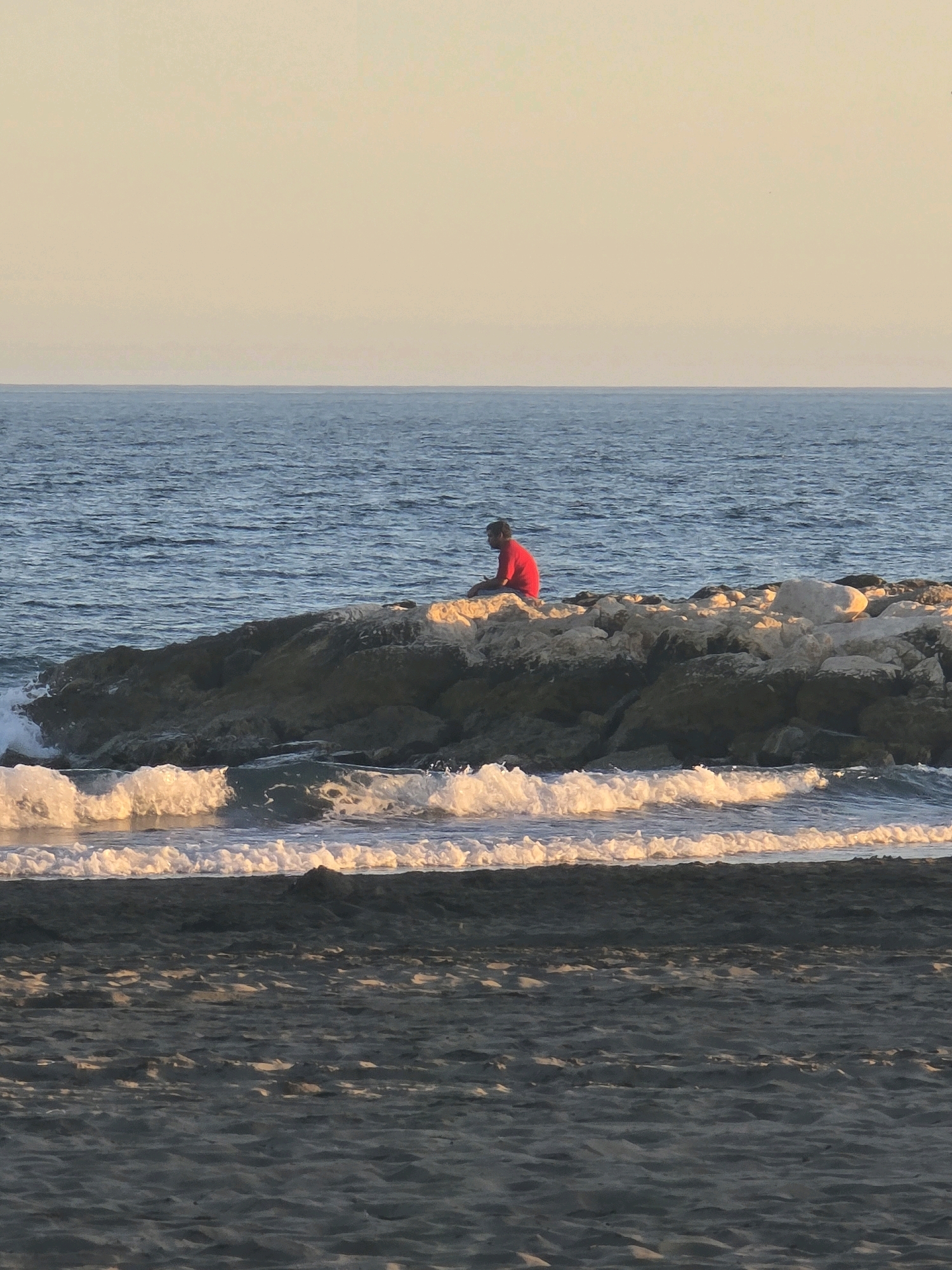 The image captures a person in a red shirt sitting on a rocky outcrop at a beach in Málaga, Spain. The person is facing away from the camera, towards the ocean. The sea is calm, with gentle waves lapping at the shore. The sky is a pale, hazy orange, suggesting either early morning or late afternoon. The beach itself is sandy, with a dark, wet appearance closer to the water, indicating recent tide activity. The rocky outcrop is made up of rough, irregular stones, with some lighter-colored rocks visible. The overall atmosphere is one of solitude and quiet contemplation.