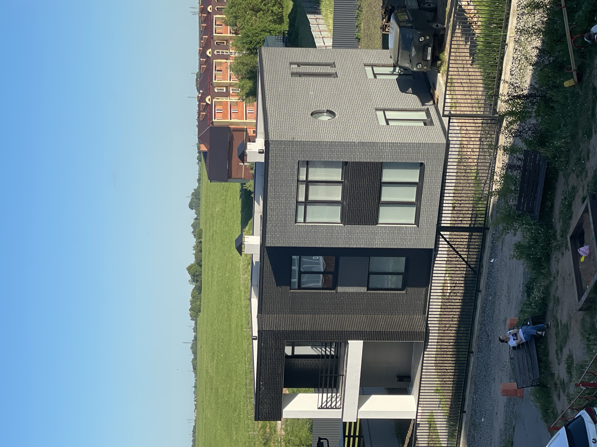 The image depicts a contemporary two-story house with a flat roof, situated at the edge of an urbanized area bordering an expansive green field under a clear blue sky. The main house features a mixed facade: the left section is white with dark, vertically slatted panels and includes an upper-level balcony with a black metal railing, while the larger right section is constructed from dark grey brick with prominent rectangular dark-framed windows and a single circular window on the upper right. A black-capped white chimney is visible on the roof.
Behind the main house, a row of terracotta-roofed, multi-story residential buildings with light-colored facades and distinct arched window designs extends into the mid-ground. Further back, an open green field stretches to the horizon, dotted with trees and power transmission lines.
In the immediate foreground, a black metal picket fence separates the main property from a semi-paved, gravelly area with patches of grass. A single individual, appearing to be a woman, is seated on a dark wooden bench, wearing a white t-shirt and blue jeans, holding an object to her mouth. To her right, another identical empty bench is visible, along with a wooden sandpit and a small playground component (seesaw). The front portion of a white car is visible in the bottom left corner.
Within the fenced property of the main house, a dark green, older model utility truck (UAZ-452 type) is parked, displaying a Russian license plate "W538MA 35". The "35" regional code identifies the location as Vologda Oblast, Russia. The scene is illuminated by bright daylight, casting distinct shadows.