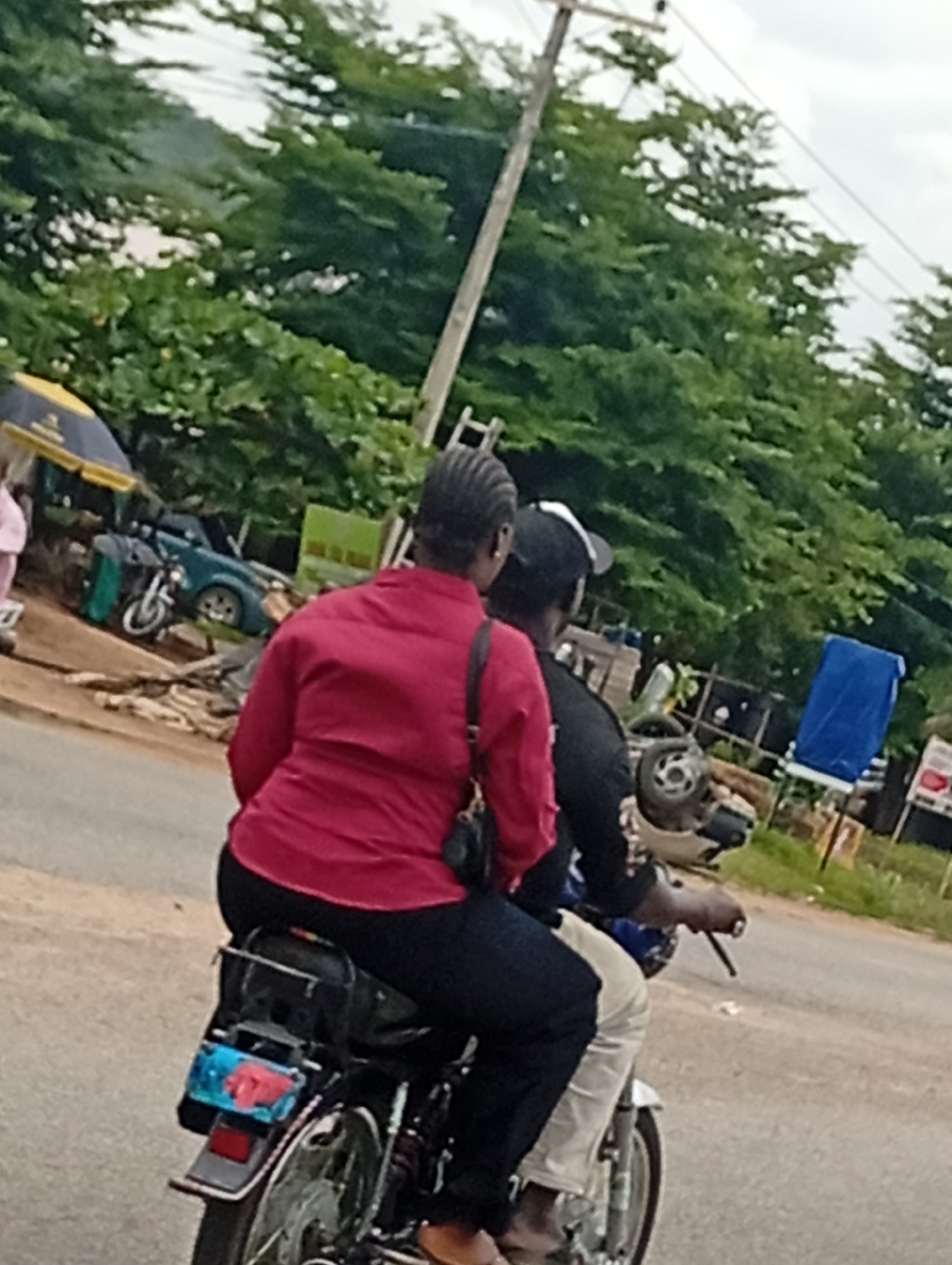 Two individuals, a woman and a man, are riding on a motorcycle on a paved road. The woman is seated behind the man, wearing a red long-sleeved shirt and black pants, with her hair in braids. The man, wearing a black shirt and a black baseball cap, is operating the motorcycle. In the background, there are lush green trees and a utility pole. To the left of the motorcycle, a blue tarp is visible, and further back, a green structure and part of a car can be seen. The overall scene suggests a typical street view in Adankolo, Nigeria, under an overcast sky.