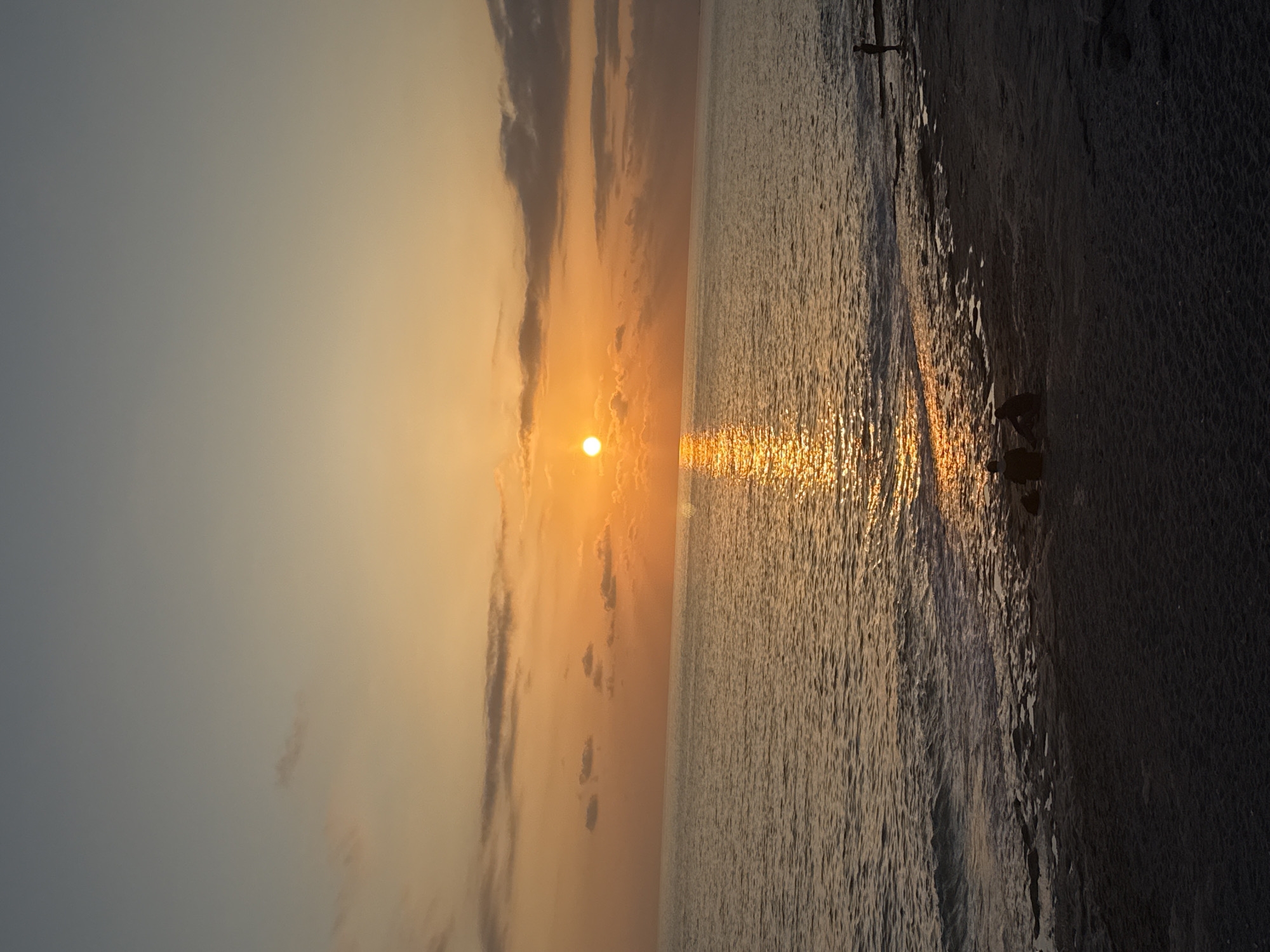 The image captures a serene sunrise or sunset over a body of water, likely an ocean or large lake, as viewed from a beach. The bright, golden-yellow sun is positioned low on the horizon towards the left, its intense light creating a shimmering, golden path that stretches across the water's surface towards the foreground.
The sky transitions from a lighter, hazy blue-grey at the top to warm orange and yellow tones near the horizon. Several elongated, dark clouds are scattered across the sky, particularly above and around the sun, adding depth to the atmospheric display.
On the right side of the frame, a dark beach or shoreline is visible, contrasting sharply with the bright water and sky due to the backlighting. Several human figures are present, appearing as dark silhouettes. One small figure stands in the distance near the horizon, to the right of the sun's reflection. Closer to the viewer, two more distinct figures are on the beach or at the water's edge; one appears to be bending or kneeling, while the other stands nearby. Other faint, dark outlines further along the beach suggest more individuals. A vertical, dark pole or structure is also visible in the upper right portion, possibly part of a pier or a marker in the water.
The scene exudes a peaceful and tranquil atmosphere, characteristic of dawn or dusk. The dominant colors are warm oranges, yellows, and golds in the sky and water reflections, set against the darker blues and grays of the upper sky and the deep silhouettes of the foreground elements. There is no visible text in the image.