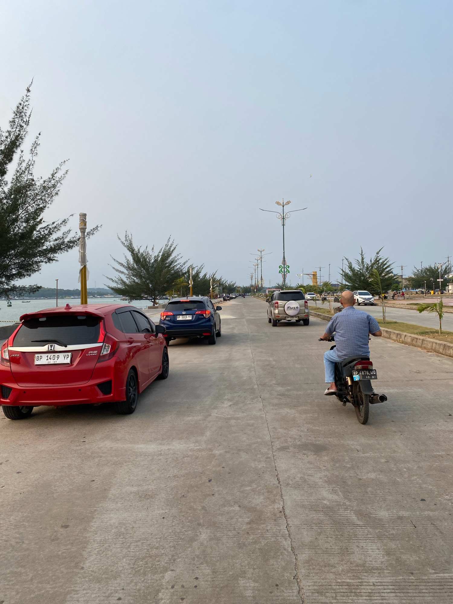 The image depicts a street scene in Tanjung Pinang, Indonesia, likely along a coastal road. The foreground features a red Honda Jazz car facing away from the viewer, parked on a concrete road. To the right of the red car, a man on a black motorcycle is riding away from the camera, wearing a blue checkered shirt and blue jeans. In the middle ground, a dark blue Honda HR-V is driving away, followed by a silver Toyota Rush SUV. Further down the road, more vehicles and pedestrians are visible, with streetlights lining the median. Trees with feathery foliage are present on the left side of the road, and the sky is overcast. In the background, a body of water can be seen, suggesting a proximity to the coast.