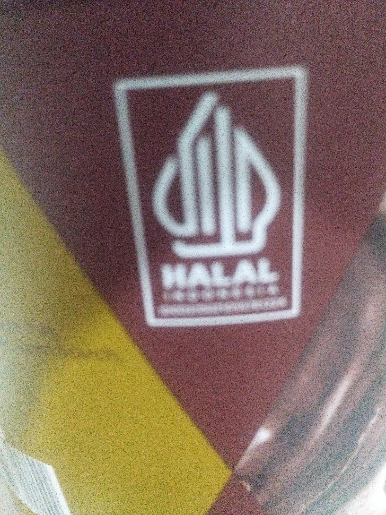 This image is a close-up of a product package with a Halal certification logo. The background is a mottled burgundy color. The logo itself is a white outline enclosing a stylized symbol and text. The symbol appears to be an abstract design with several vertical elements. Below the symbol, the word "HALAL" is prominently displayed in white capital letters. Beneath "HALAL," there is smaller text, likely indicating the certifying body and location, which appears to be "INDONESIA."
The overall image has a grainy texture, suggesting it may have been taken with a low-resolution camera or in poor lighting conditions. The lighting seems to be coming from the right, casting a slight shadow or reflection on the right side of the image. A diagonal band of yellow color is visible on the left side of the package, creating a visual contrast with the burgundy background. This yellow area has some indistinct text or pattern on it, but it is too blurry to be legible. The image focuses on the Halal certification, which is a significant detail for products intended for consumption by Muslims. Given the location context of Babelan, Indonesia, the presence of an Indonesian Halal certification is highly relevant. There are no people or active events depicted; the image is a static shot of product packaging.