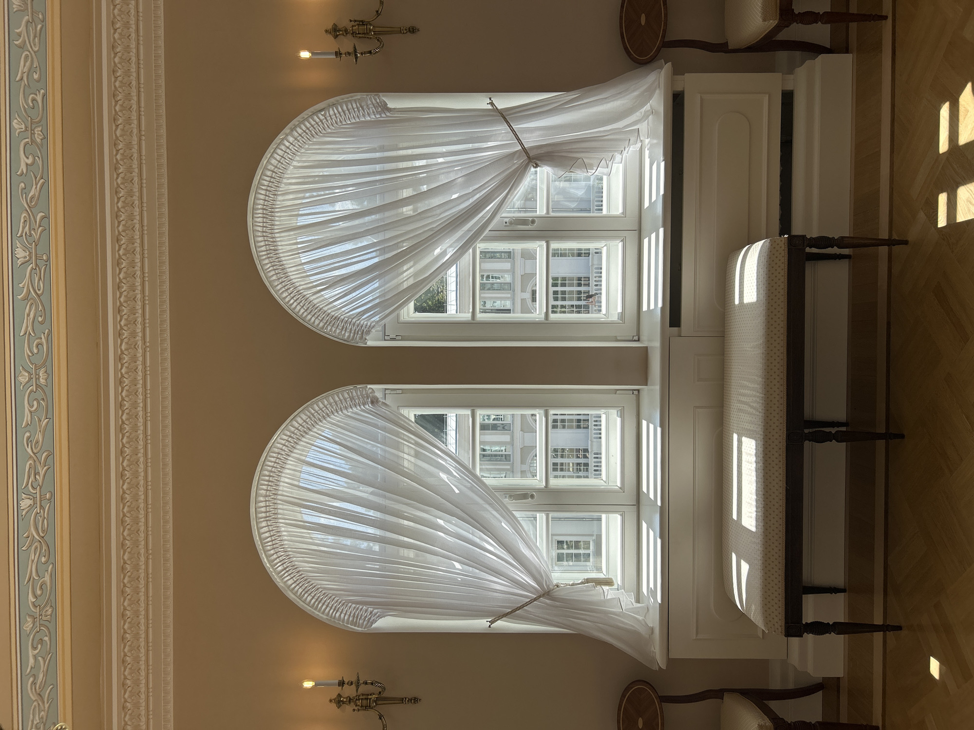 The image depicts an interior scene with two arched windows covered by sheer white curtains. Sunlight streams through the windows, casting striped patterns on the floor and a bench positioned in front of them. The bench is upholstered in a light-colored fabric with a subtle pattern and is supported by dark wooden legs. On either side of the bench, partially visible, are two chairs with dark wooden frames and light-colored upholstered seats.
The walls are painted a light, warm color. Above the windows, ornate decorative moldings are visible on the ceiling, with a band of pale blue and gold detailing. Two wall-mounted sconces, each with a single lit candle, are positioned on the walls to the left and right of the windows, adding to the soft lighting of the room.
Through the windows, a building with multiple floors and windows is visible, suggesting an external view of architecture. The floor is a polished wooden parquet. The overall impression is one of elegance and tranquility. The location is identified as the city of Dobasnenskі Selski Savaet, Belarus.