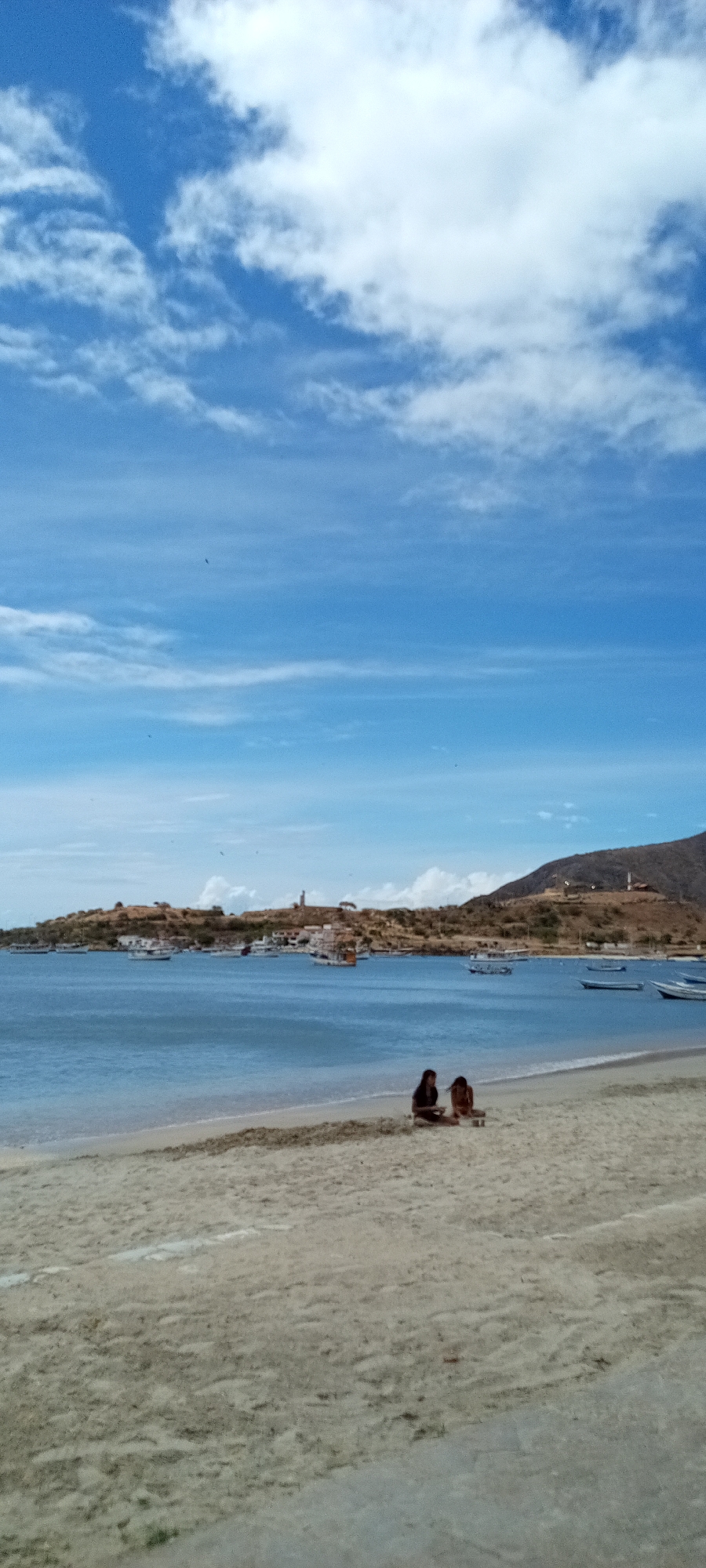 The image shows a beach scene on a sunny day. In the foreground is a sandy beach with two people sitting on it. Behind them is a body of water, likely the sea or a bay, with several boats visible. In the background, there's land with some buildings and what appears to be a hill or mountain. The sky is blue with scattered white clouds. The location is Unknown.