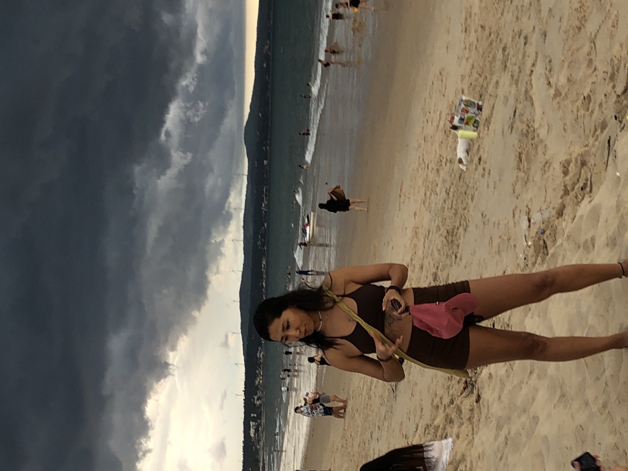 This image captures a beach scene in Phu Thuy Ward, Vietnam, under a dramatic, cloudy sky. In the foreground, a woman with dark hair stands on the sandy beach, wearing a dark bikini top and brown shorts. She has a yellow strap across her body and is holding a pink cap and her phone. Behind her, the ocean is visible with waves breaking on the shore and several people wading and swimming in the water. Further in the distance, a coastline with hills and wind turbines can be seen. The beach is populated with various individuals, some standing and interacting, others walking along the water's edge. The overall atmosphere suggests a somewhat overcast day at the beach.