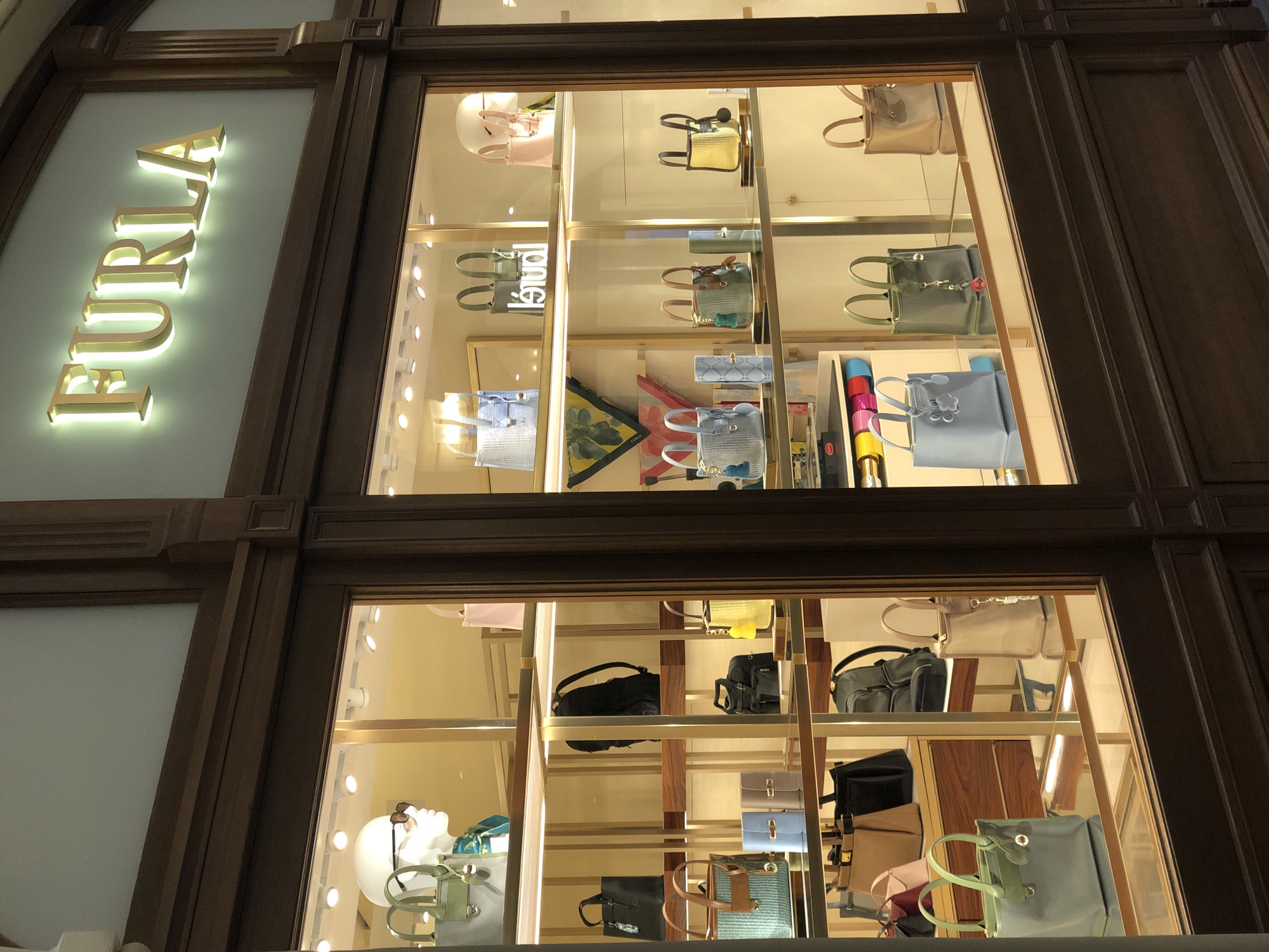 A well-lit display window of a Furla store presents an array of handbags. The window is part of a dark wood-framed facade. The Furla sign is illuminated in a bright golden light on the left side of the window. The display features multiple shelves filled with various styles and colors of handbags, some arranged individually, others in groupings. Mannequin heads with accessories are visible on the lower shelves.
Given the architecture, interior design, and the Furla brand, the location appears to be in an upscale shopping district, possibly in Moscow, Russia.