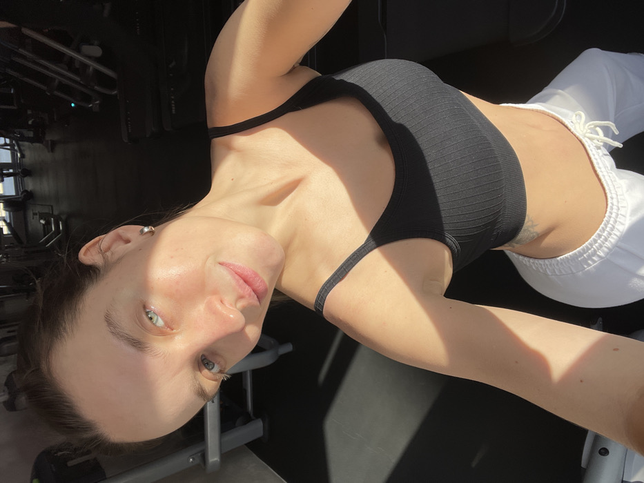 A light-skinned adult female with light-colored eyes, dark hair pulled back, visible freckles, and a small silver hoop earring in her left ear is depicted in an indoor gymnasium. She wears a black ribbed sports bra and white drawstring sweatpants; a tattoo is visible on her lower left abdomen. The subject is taking a selfie, looking directly at the camera with a slight, closed-lip smile. The background features dark-toned gym equipment, including exercise machines and treadmills, on a dark floor. Direct natural light illuminates the subject from the left, creating defined highlights and shadows across her face and body. The precise location within Budva, Montenegro, is not discernible from the visual information provided.