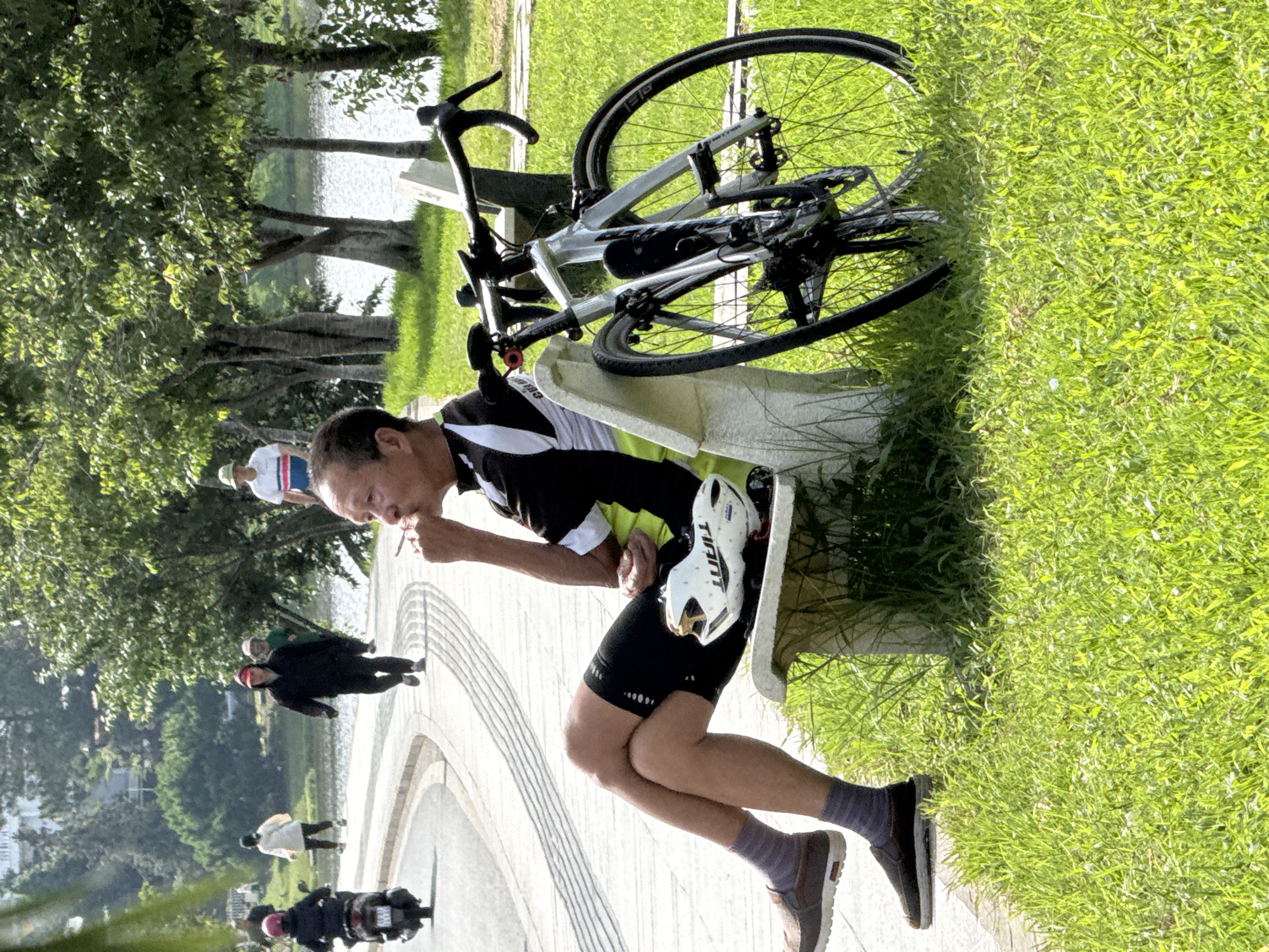 A man sits on a park bench in Da Lat, Vietnam, with a bicycle parked beside him. The man, wearing cycling attire and socks, holds a cigarette to his lips. A white helmet rests on the bench next to him. The scene is set beside a pathway and a body of water, with trees and other park-goers visible in the background. The sun casts shadows, indicating it is daytime.