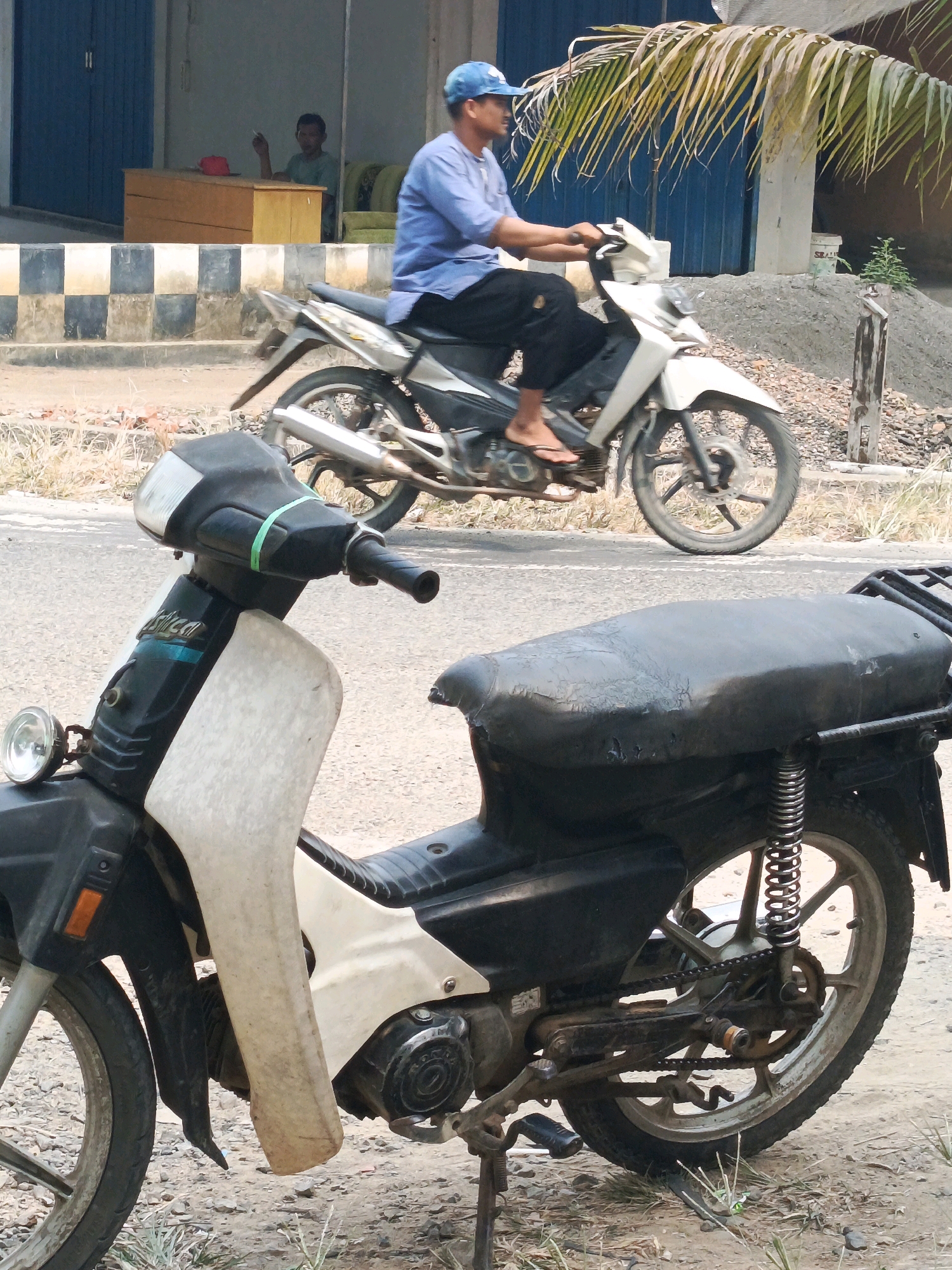 A middle-aged man wearing a blue shirt and a blue cap rides a white and black motorcycle. He is wearing flip-flops and black pants. The motorcycle is parked on the side of a dirt road. In the background, there is a building with a blue door and a wooden desk. A person is sitting behind the desk. The image appears to have been taken during the day, with bright sunlight casting shadows. The environment looks like a street in a rural or semi-urban area in Wonosari, Indonesia.