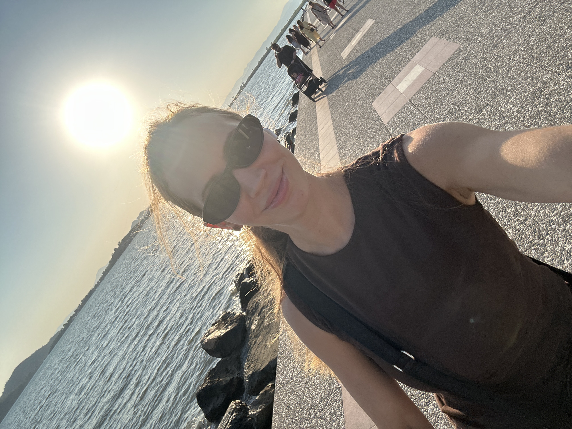 A woman with blonde hair, wearing sunglasses and a brown tank top, takes a selfie on a paved promenade next to the sea. The sun is bright in the sky, casting a warm glow. In the background, several people are walking along the promenade, some pushing strollers. The sea is calm with a rocky shoreline to the left. Mountains are visible in the distance. The overall scene suggests a pleasant day in Fethiye, Turkey, possibly in the late afternoon.