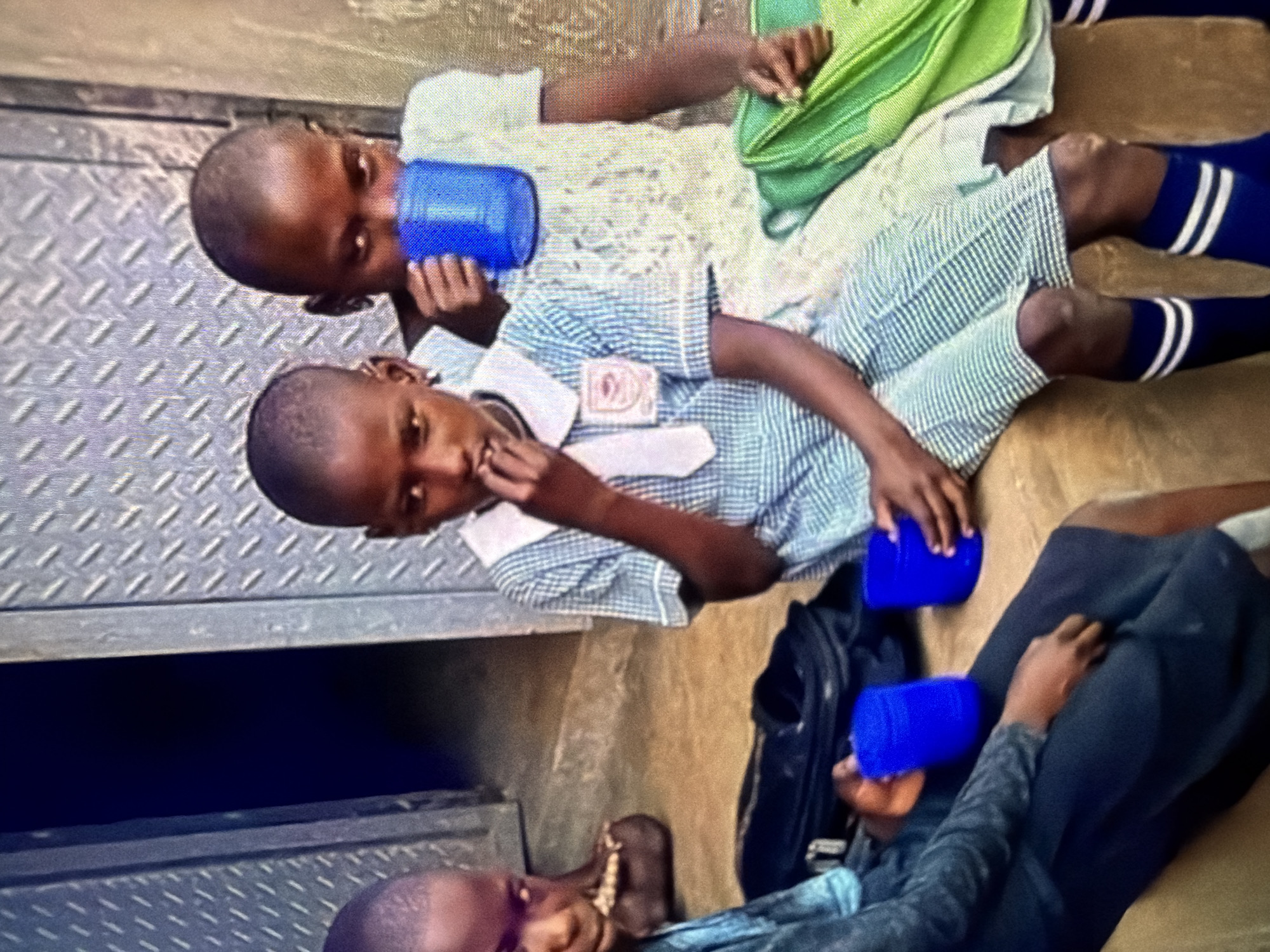 Four young children are seated outdoors, likely on the ground, in front of a corrugated metal wall and a doorway. The child in the center, wearing a blue and white checkered school uniform with a white collar and tie, is bringing a finger to their mouth. To their right, another child, wearing a lighter colored garment, is holding a blue cup to their lips. To the left of the child in the uniform, another child, wearing a dark blue garment, is holding a blue cup. The furthest child on the left is partially visible, wearing a dark blue outfit. Green backpacks are visible on the right side of the image, and a dark bag is on the ground in front of the children. The environment appears to be a simple, possibly urban, setting with a concrete floor.
