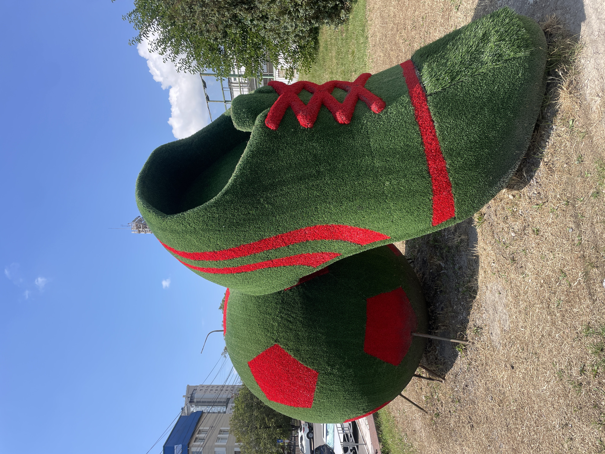 This is a medium shot, eye-level, outdoor, daytime photograph of a large, green, artificial grass sculpture of a soccer ball and a soccer cleat. The sculpture is located in Novorossiysk, Russia. The soccer ball is on the left side of the image, and the soccer cleat is to its right and slightly behind it. Both are decorated with red accents. The soccer ball has red pentagons and hexagons, mimicking a traditional soccer ball pattern. The soccer cleat has red stripes along its side and red laces.
The background shows a clear blue sky with a few white clouds. To the left, there are buildings, including a multi-story residential building with a blue roof and a taller, modern building. A street with cars is visible in the distance. To the right of the sculpture, there is a grassy slope with trees and a chain-link fence. The ground in front of the sculpture is covered with dry, straw-like grass and some dirt. There are no people visible in the image. The overall scene appears to be a public space, possibly a park or a decorated city area. The sculpture itself is the main subject, presented in a way that highlights its whimsical and oversized nature.
