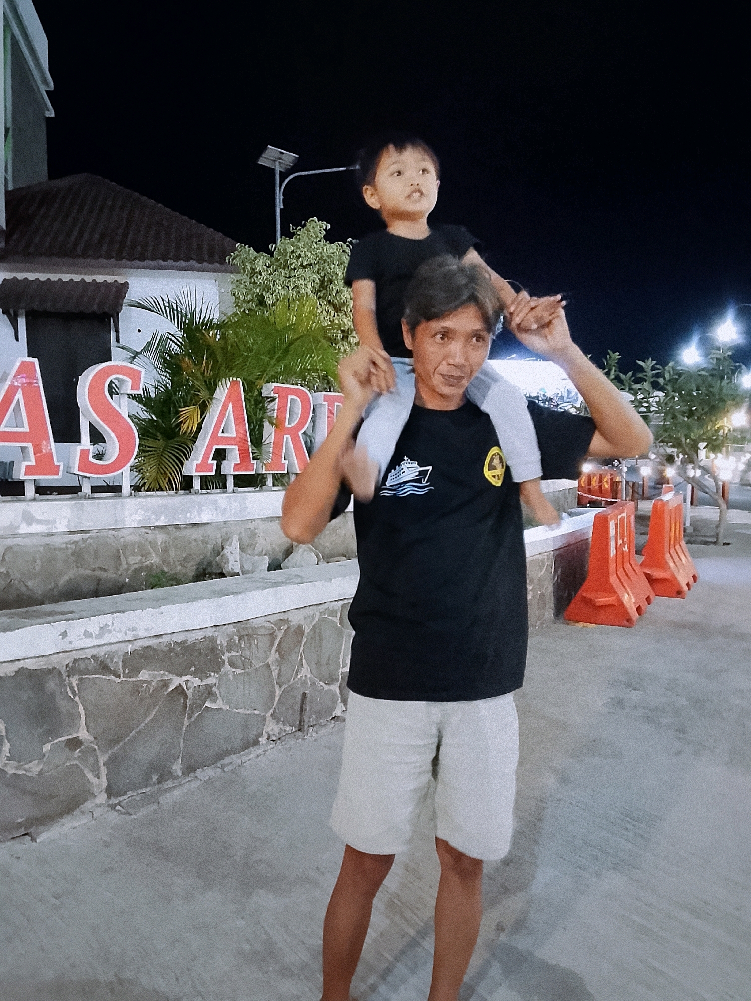 A father is carrying his son on his shoulders at night in front of a large sign that says "AS ART". The father is wearing a black t-shirt with a white boat graphic and a yellow circular logo, and white shorts. The son is wearing a black t-shirt and is looking to the left. The sign is lit with red lettering. In the background, there are trees, streetlights, and orange traffic barriers. The location appears to be an outdoor public space, possibly a street or a park in Boyolali, Indonesia. The ground is paved, and there is a stone wall behind the sign.