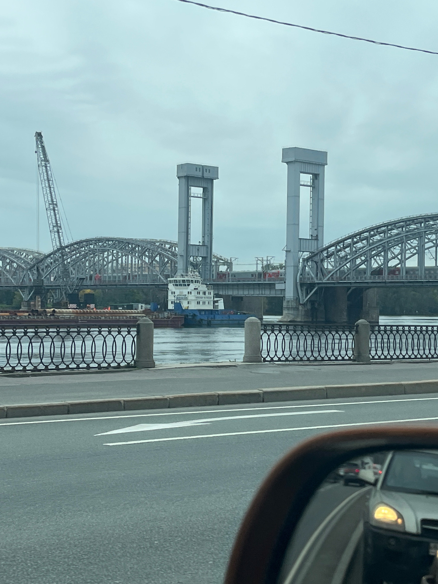 An overcast day scene in Saint Petersburg, Russia, shows a view from a vehicle. In the foreground, a car's side mirror reflects traffic on a wet road with directional arrows. Beyond the mirror, a wide river with a decorative iron fence and bollards along its bank is visible. A barge with yellow cylindrical objects is moored on the river, with a tugboat named "LATERBLAND" passing by. In the background, a large arched railway bridge with multiple spans dominates the scene. Two prominent vertical towers with rectangular openings stand on either side of a section of the bridge. A construction crane is visible to the left of the bridge. Trains are seen on the railway tracks of the bridge. The sky is a dull, cloudy gray.