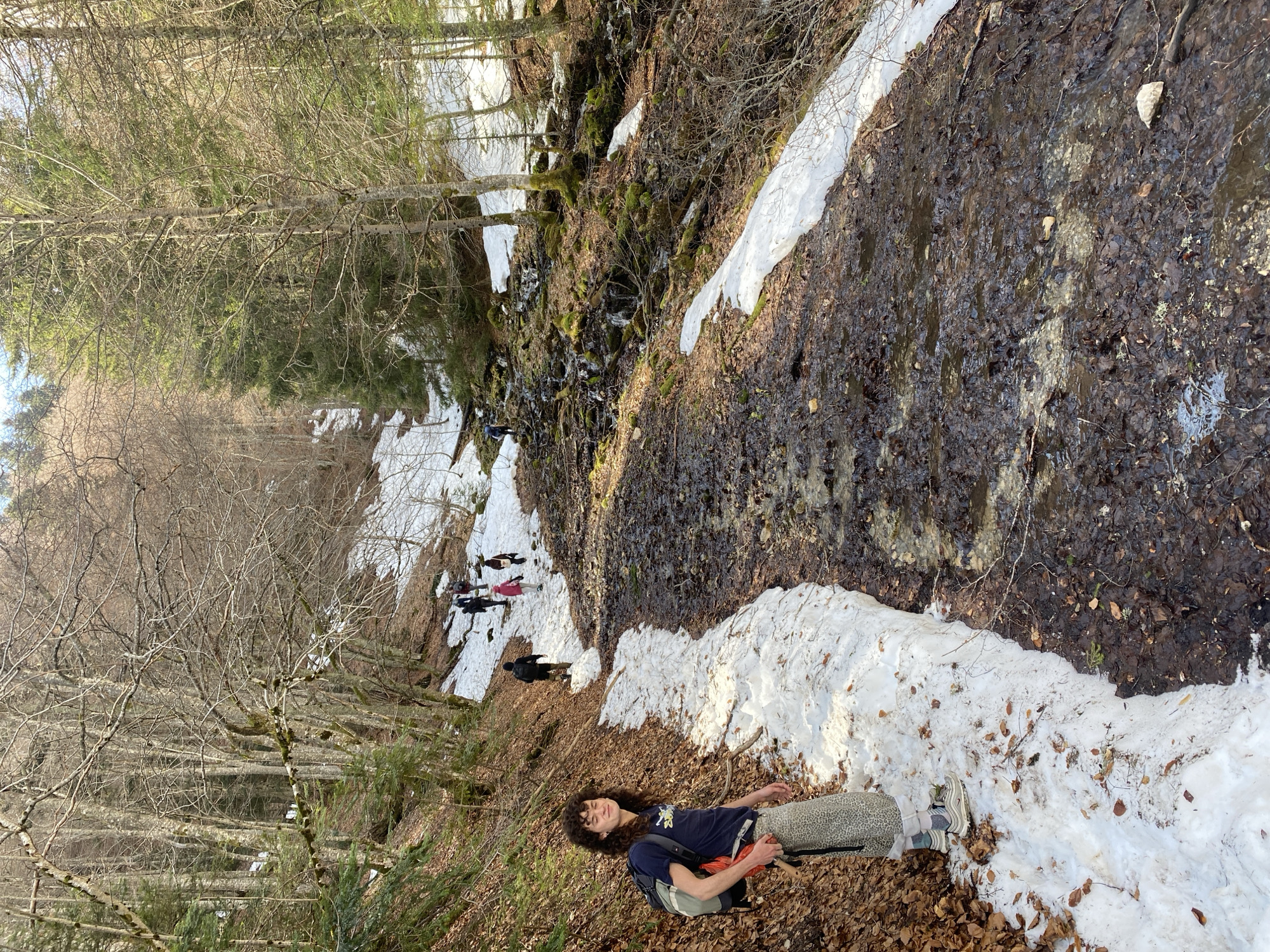 The image shows a landscape in Saint-Michel-les-Portes, France, likely in a mountainous or wooded area. The ground is covered with a mix of fallen leaves and patches of snow. A person is lying on the leaves in the foreground, wearing a dark t-shirt, patterned pants, sneakers, and carrying a backpack. A small group of people is walking along a path further up the slope. A stream or small waterfall cascades down the hillside covered in vegetation. Trees, mostly bare of leaves, are visible on both sides of the path. The overall environment suggests a late winter or early spring setting.