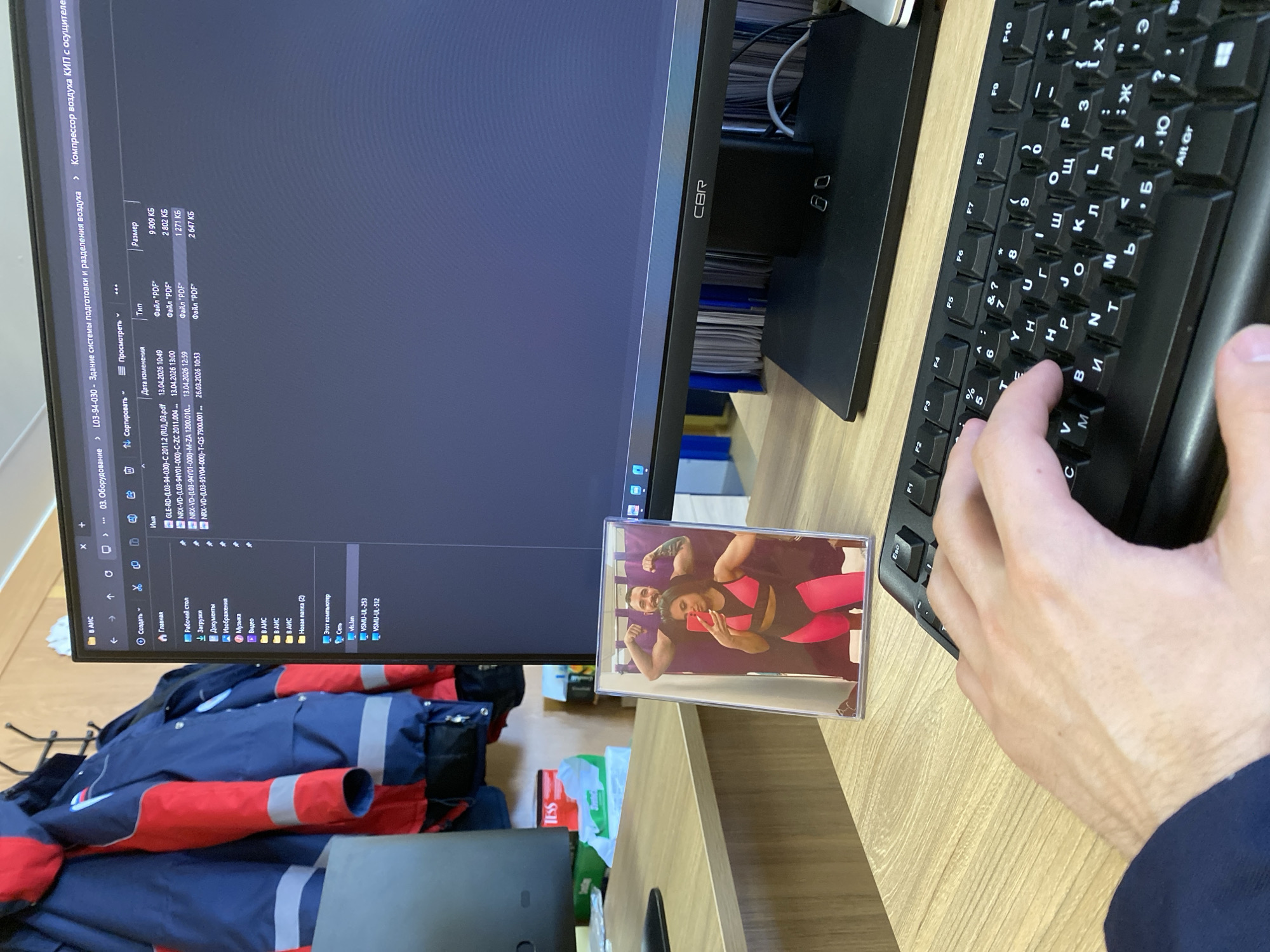 A desk setup includes a computer monitor displaying files related to a "building system for preparation and separation of air" and "KIP air compressor with dehumidifier," a black keyboard with Russian and English characters, a framed photograph of a couple in workout clothes, a partially visible hand typing on the keyboard, and a folded blue and red jacket. The photo on the desk shows a couple in workout attire posing, with the woman holding a phone up to take a selfie. The files on the screen are PDFs with names indicating component designations. There is no indication this image is located in Luzhitsy, Russia.