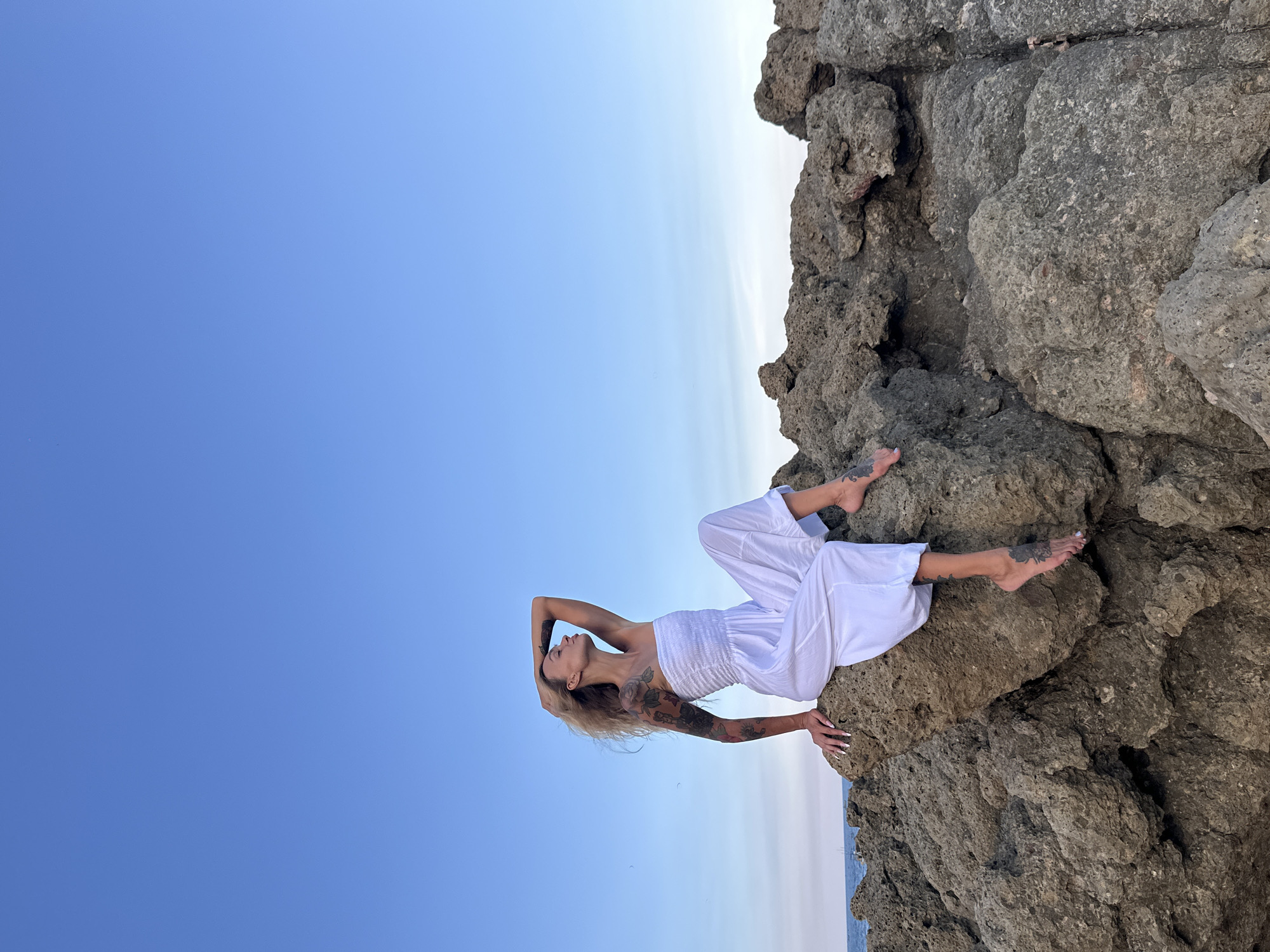A blonde woman with tattoos sits on a large rock formation near the ocean under a clear blue sky. She is wearing a white strapless jumpsuit and has her left hand raised to her head, her hair falling over her shoulder. Her bare feet are visible, one resting on the rock and the other dangling. The rocks are rough and uneven, and the ocean is visible in the background. The scene appears to be a natural coastal environment, possibly a beach or rocky coastline, but there are no specific urban elements or identifiable landmarks to confirm a location in Mũi Né, Vietnam.